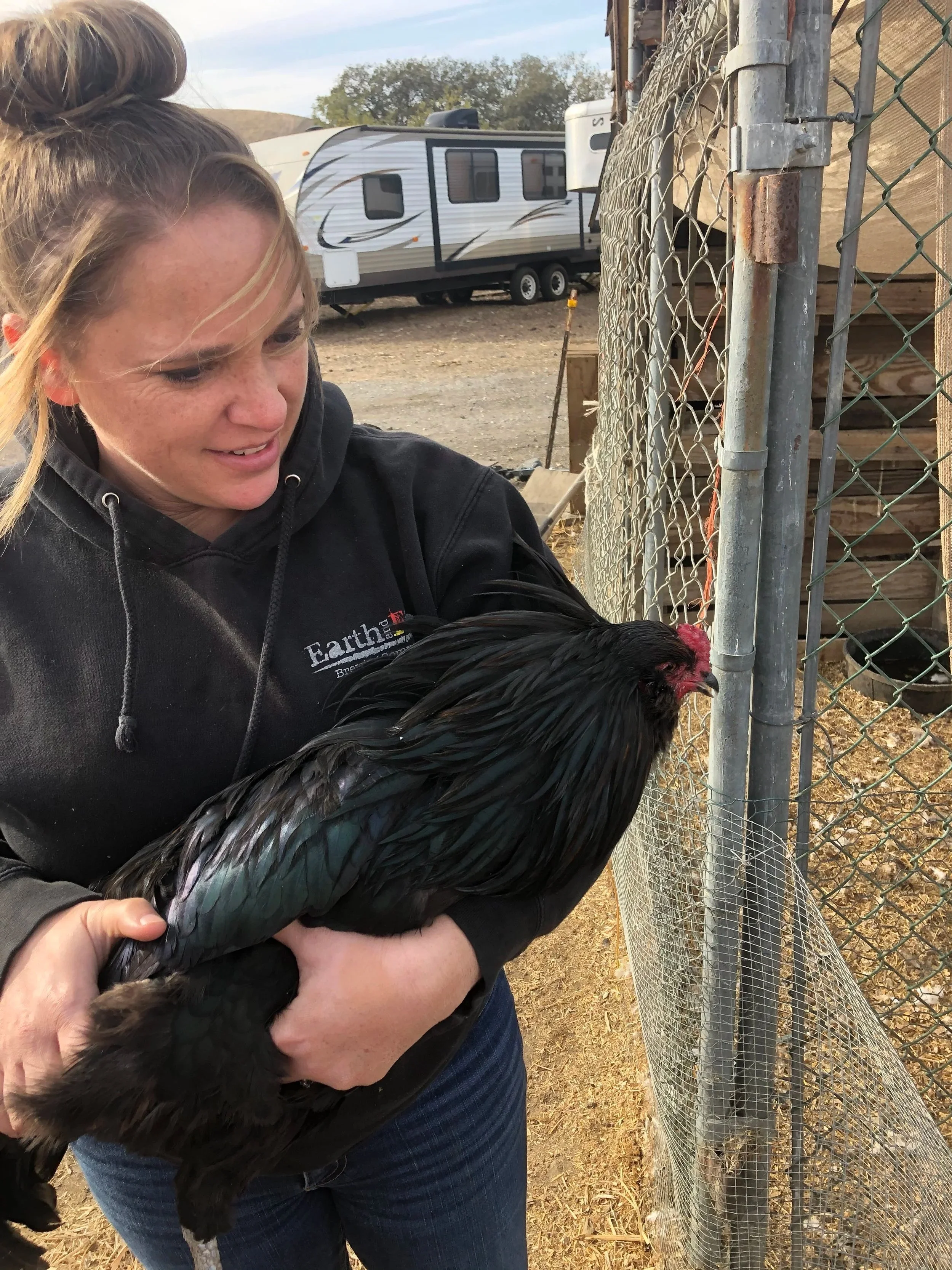 A woman holding a black rooster outside near a chain-link fence with a trailer in the background.