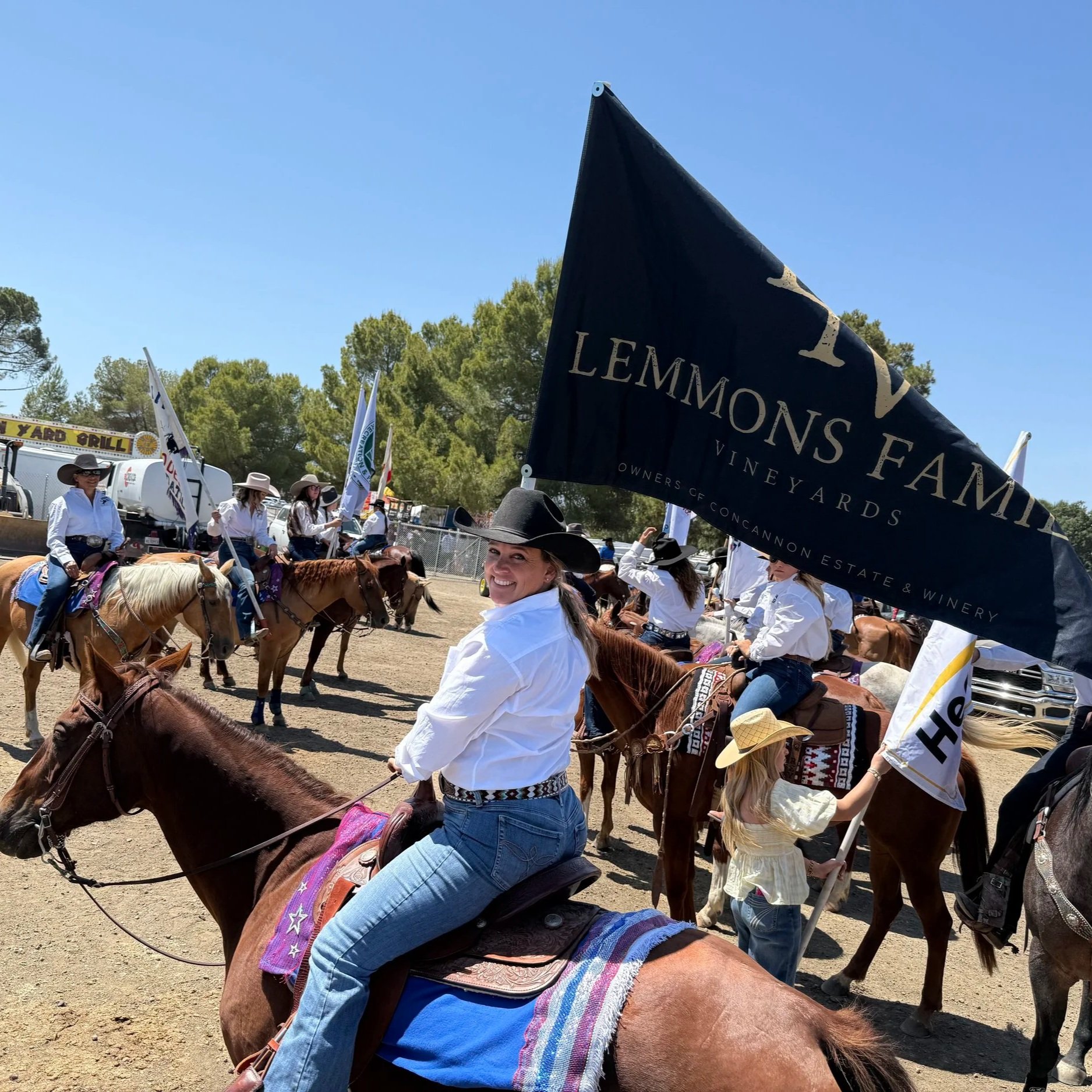 People riding and gathered with horses at a daytime outdoor event with flags and trees in the background.