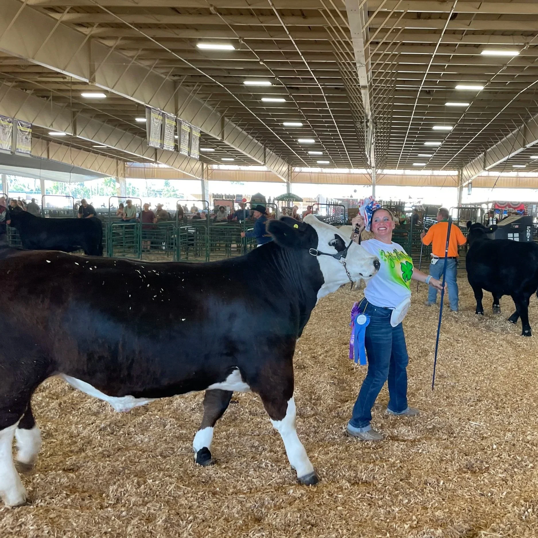 A woman holding a pole and wearing a white t-shirt with green graphics stands next to a black and white cow at a livestock show inside a large barn. The woman is smiling and the barn is filled with other animals, people, and pens in the background.