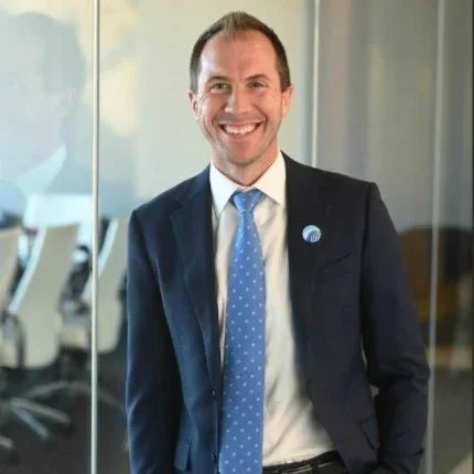 A smiling man in a business suit standing in an office with glass walls.