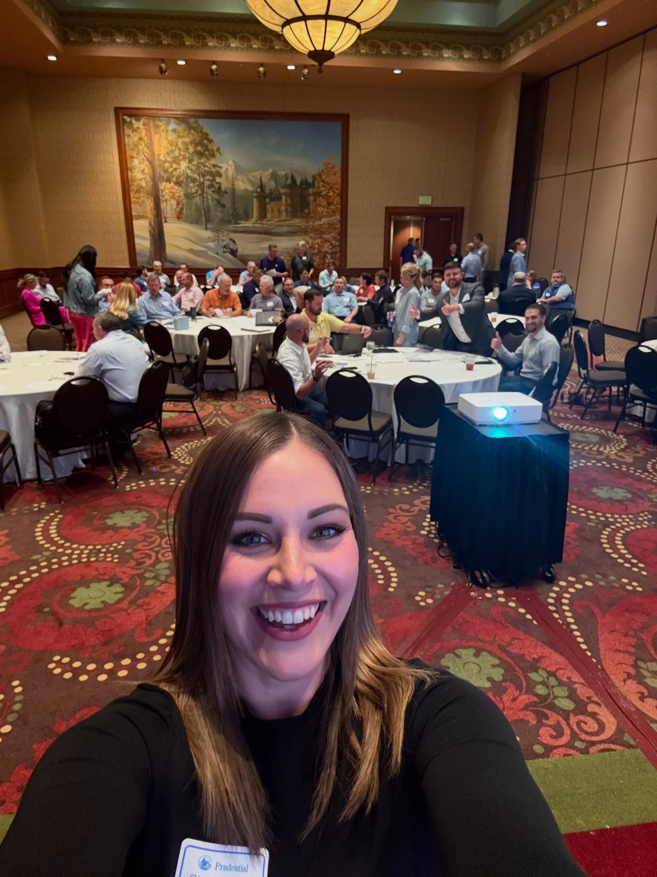 A woman taking a selfie in a large banquet room filled with round tables and many people. There is a large landscape painting of mountains and trees on the wall behind her, and a projector on a stand nearby. The woman has long brown hair, is smiling, and wearing a black top with a name tag.