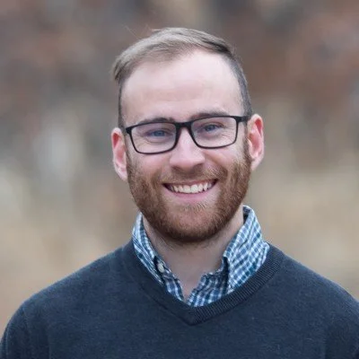 A young man with glasses, a beard, and short hair, smiling outdoors, wearing a blue sweater over a checkered shirt, with a blurred natural background.