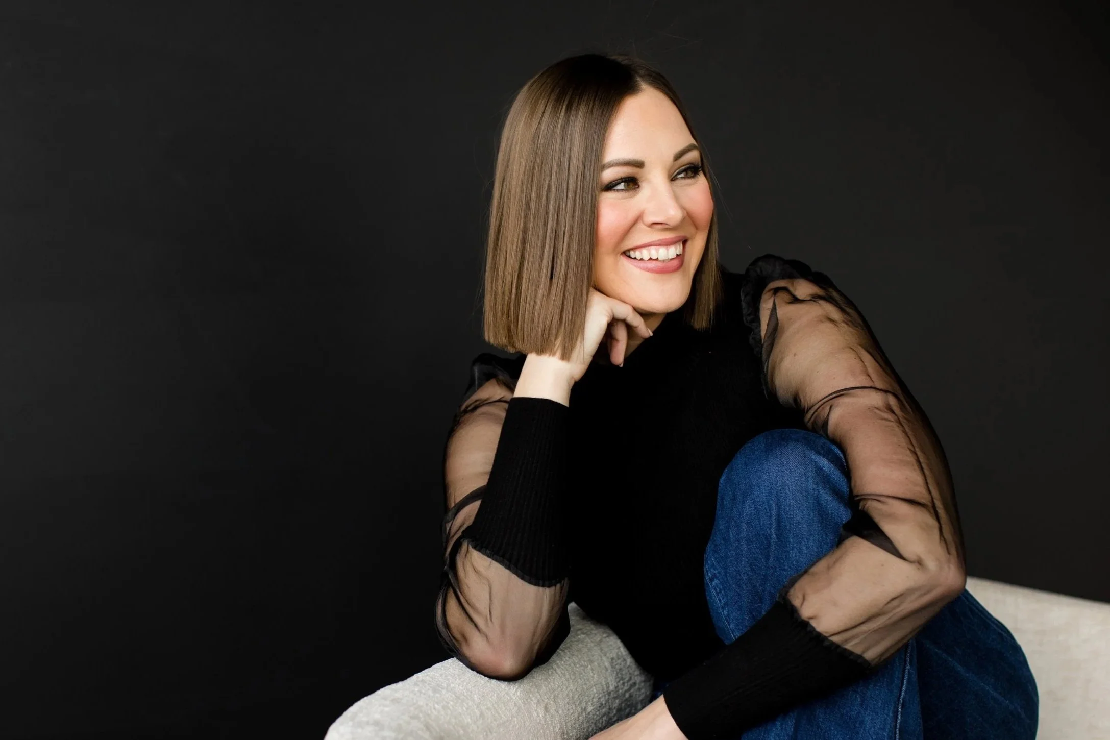 A woman with a shoulder-length brown bob haircut and a big smile, sitting on a beige couch against a black background, wearing a black top with sheer long sleeves and blue jeans.