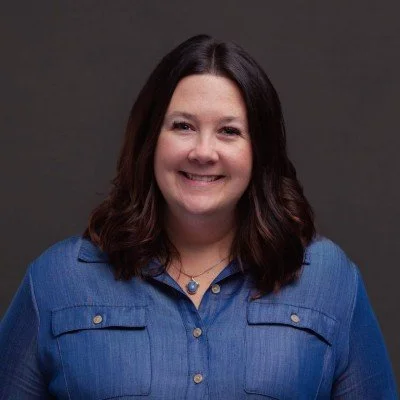 A woman with shoulder-length dark brown hair smiling in front of a gray background, wearing a blue button-up shirt.