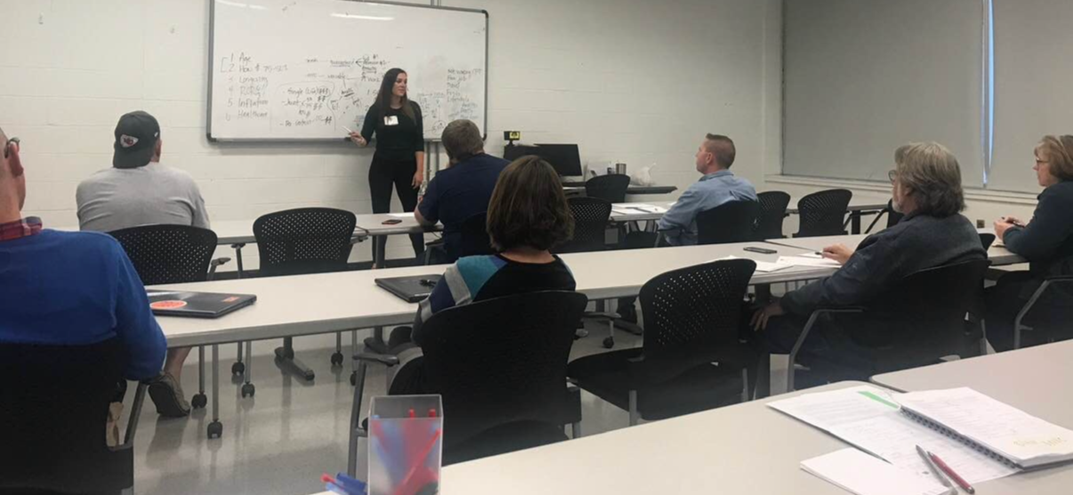 A woman standing at the front of a classroom, giving a presentation to six seated adults. The classroom has a whiteboard filled with notes and a window with closed blinds.