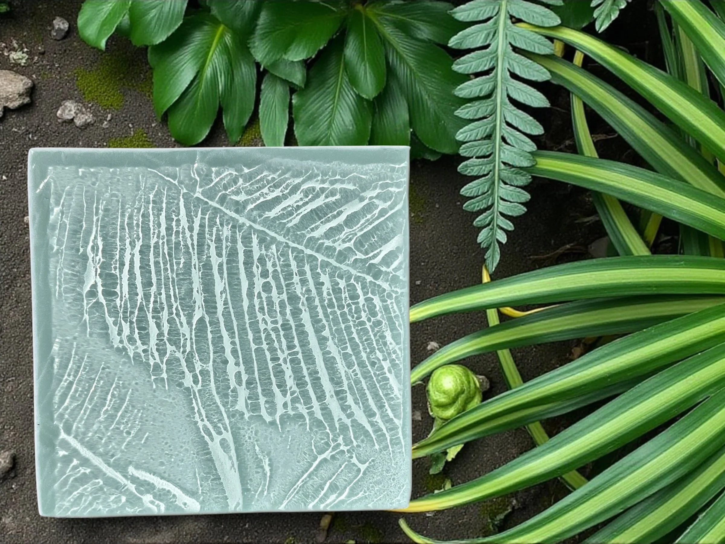 Close-up of various green tropical leaves and a small green fruit or seed on the dark soil ground, with a translucent decorative glass panel with leaf pattern in the foreground.