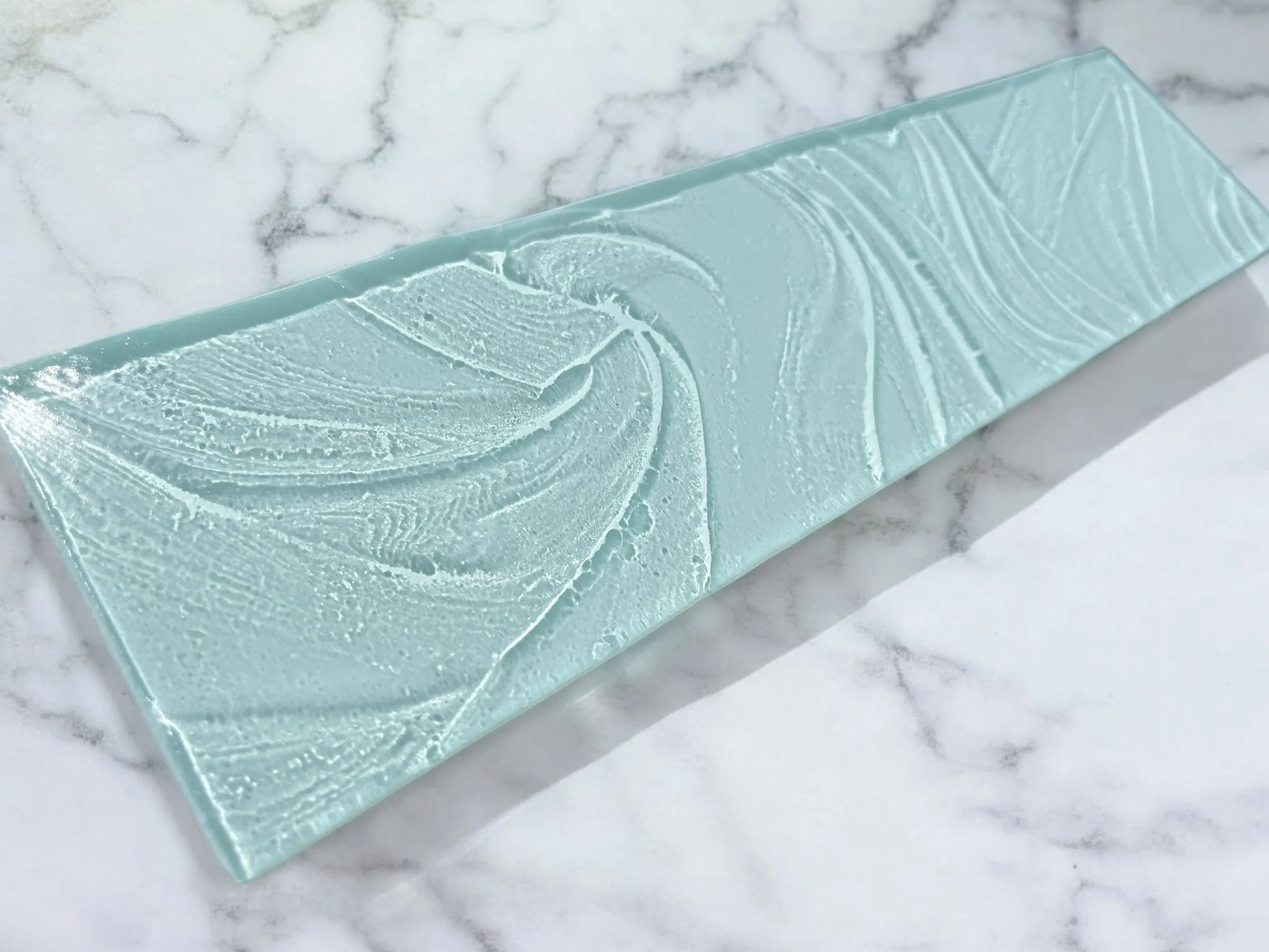 A rectangular piece of light blue-green glass with swirling textured patterns, placed on a white marble surface with gray veining.