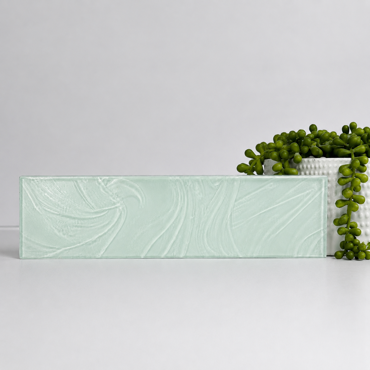 A rectangular light green marble slab is placed on a white surface, with a white textured pot filled with a trailing green succulent plant to the right.