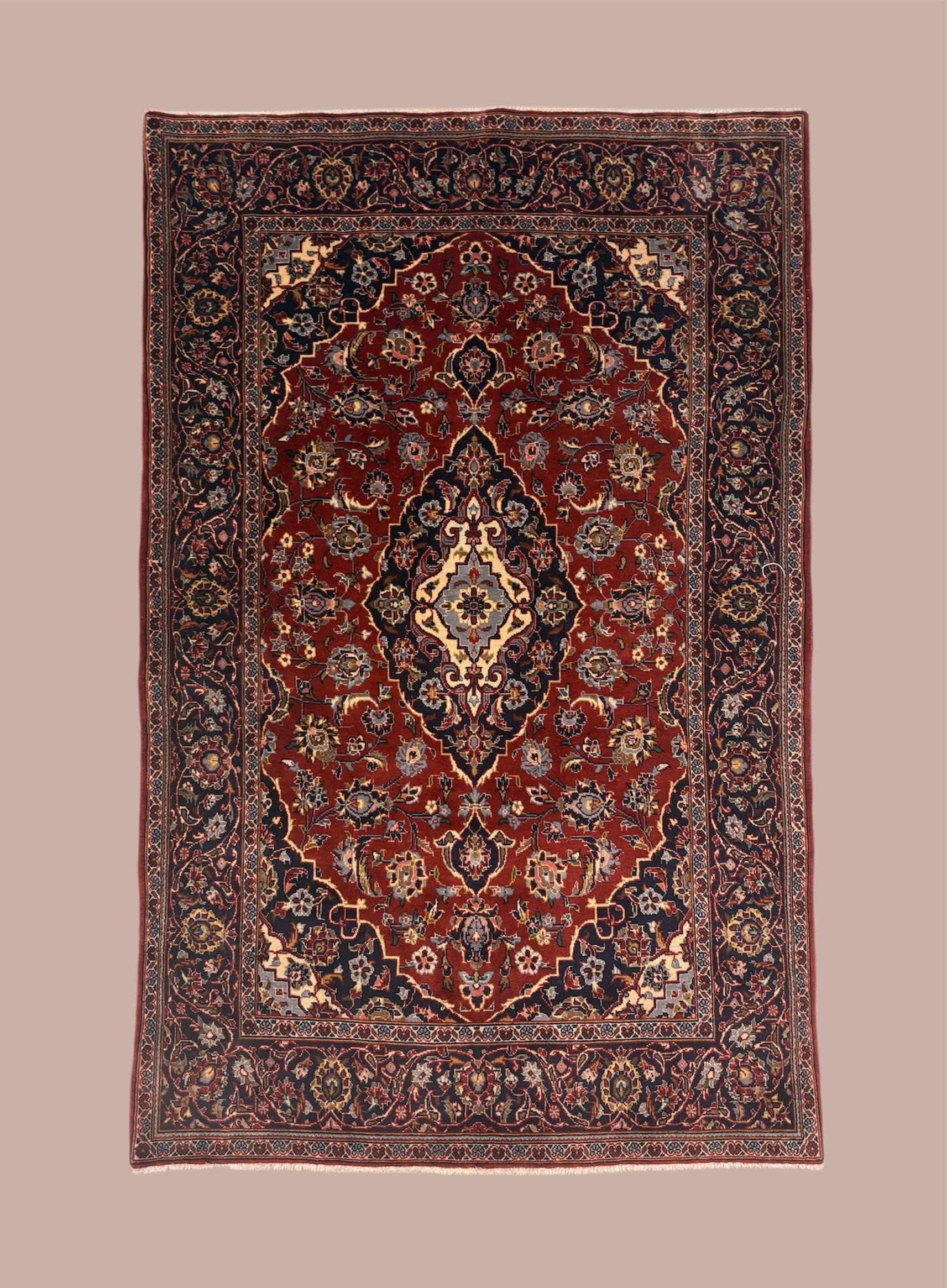 An intricately patterned Persian rug with a central floral motif, surrounded by detailed borders in shades of red, black, beige, and blue, placed on a plain beige background.