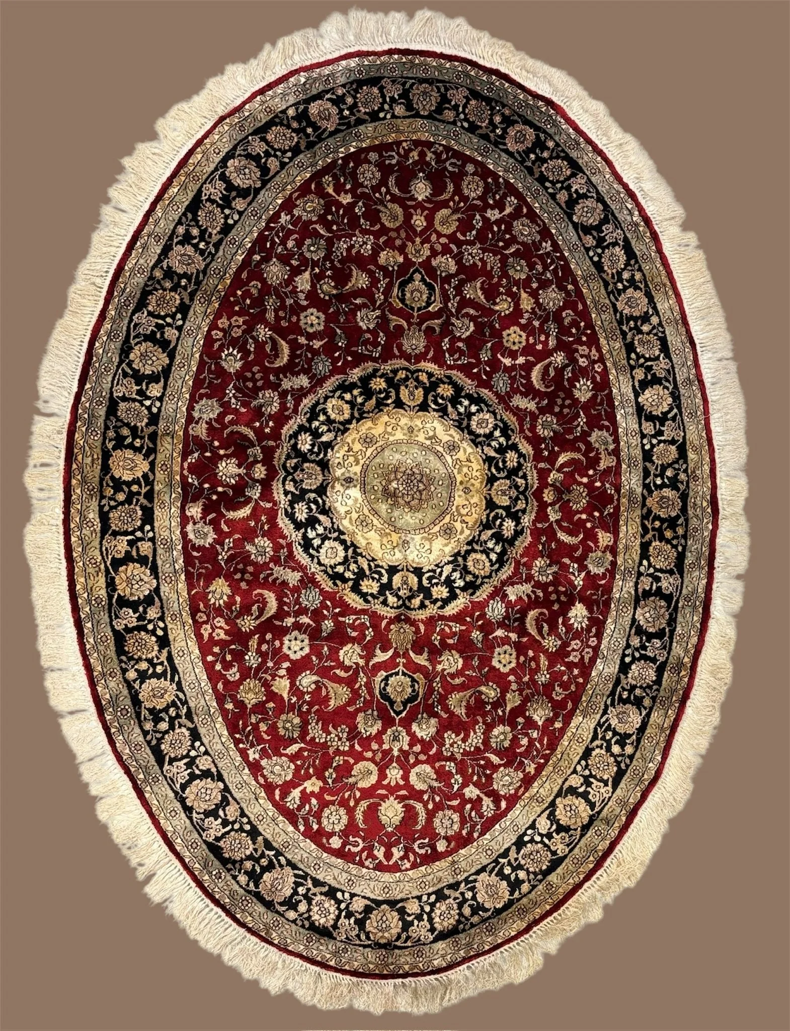 A traditional Persian-style oval rug with intricate floral patterns in red, black, cream, and beige colors and fringed edges.