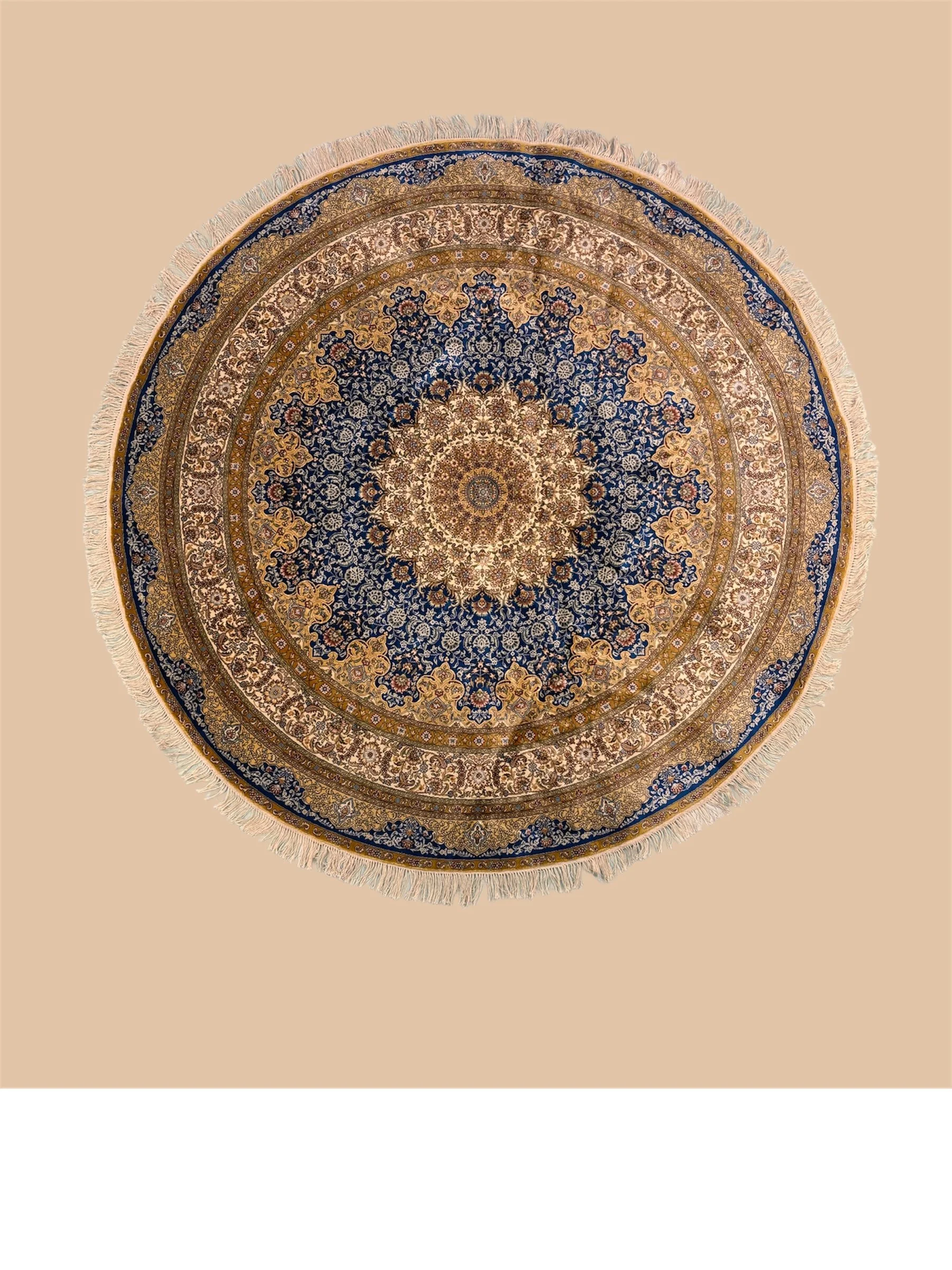 Circular ornate Persian-style rug with intricate floral and geometric patterns in blue, beige, and gold, fringed edges, placed on a plain light brown background.