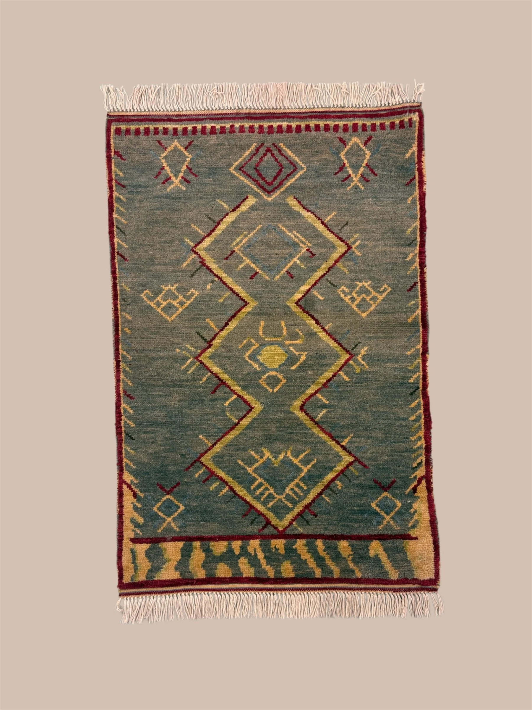A rectangular area rug with geometric patterns in shades of green, red, yellow, and blue, with fringe on the top and bottom edges, placed on a beige background.