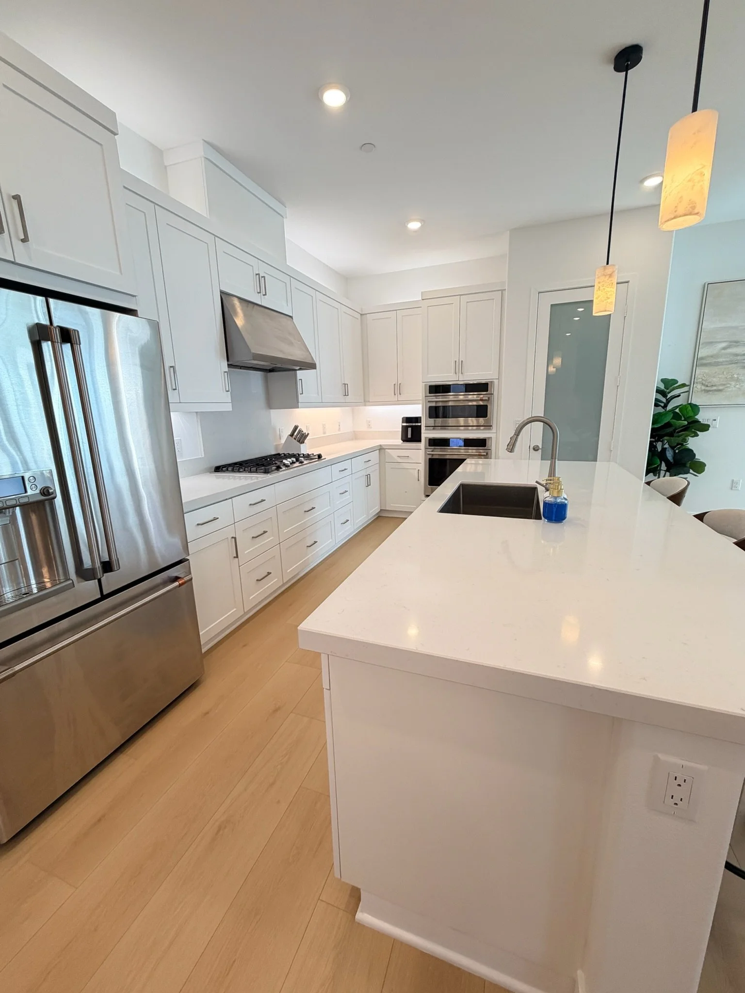 Modern kitchen with white cabinets, a large white island, stainless steel appliances, a built-in oven, a black sink with a gold faucet, pendant lights, and hardwood floors.