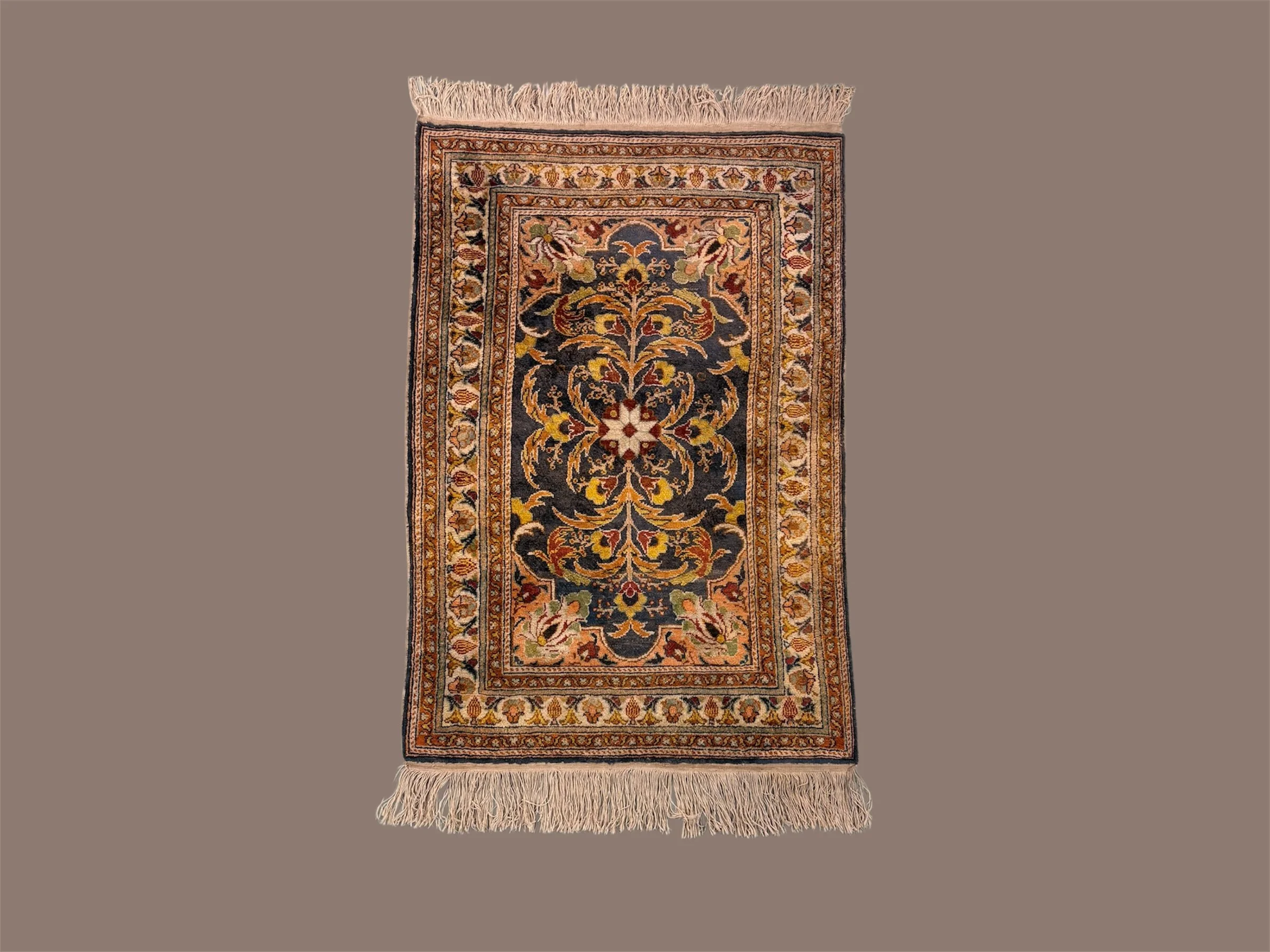A decorative area rug with floral and vine patterns in shades of black, red, yellow, green, beige, and white, bordered by multiple intricate borders, and fringes at each end, placed on a neutral surface.