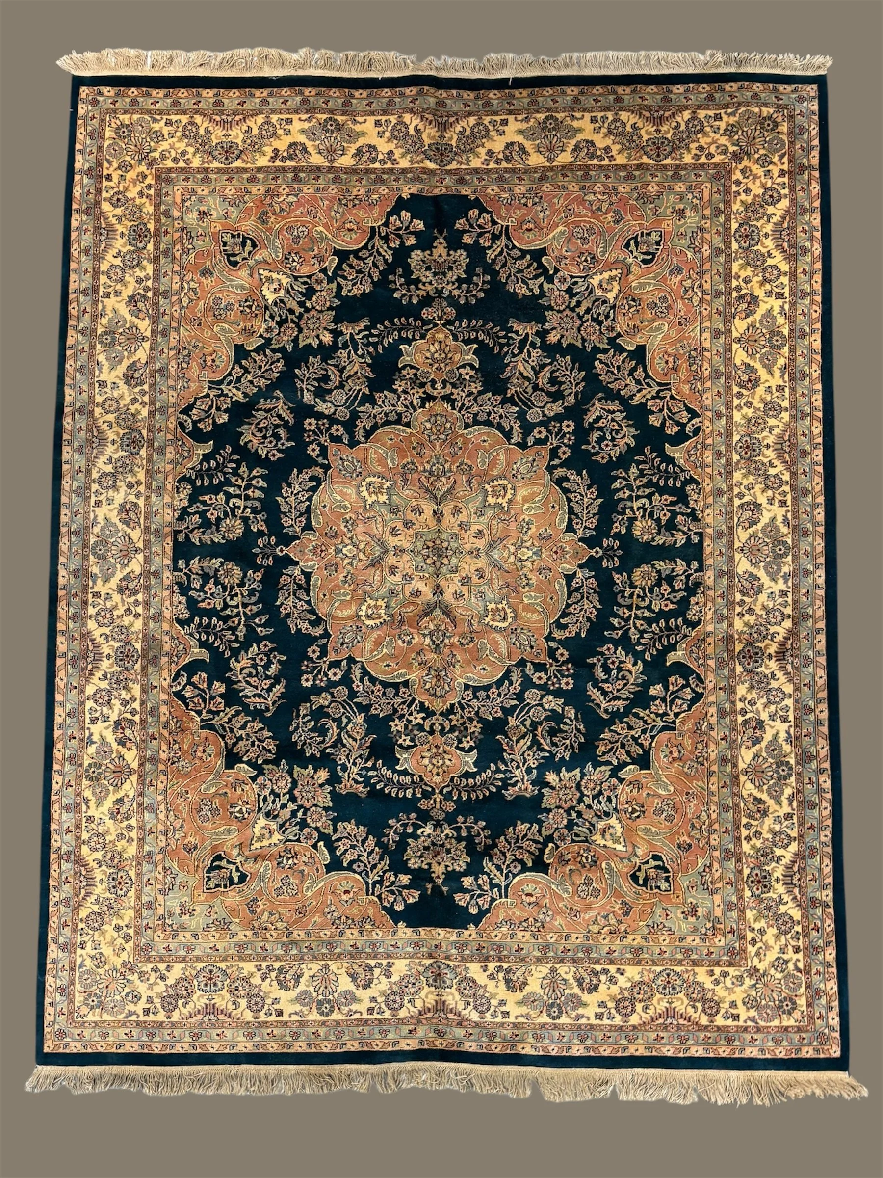 A traditional Persian-style area rug with a central floral medallion, surrounded by intricate floral and vine patterns in navy blue, beige, and peach tones, with a detailed border and fringed edges.