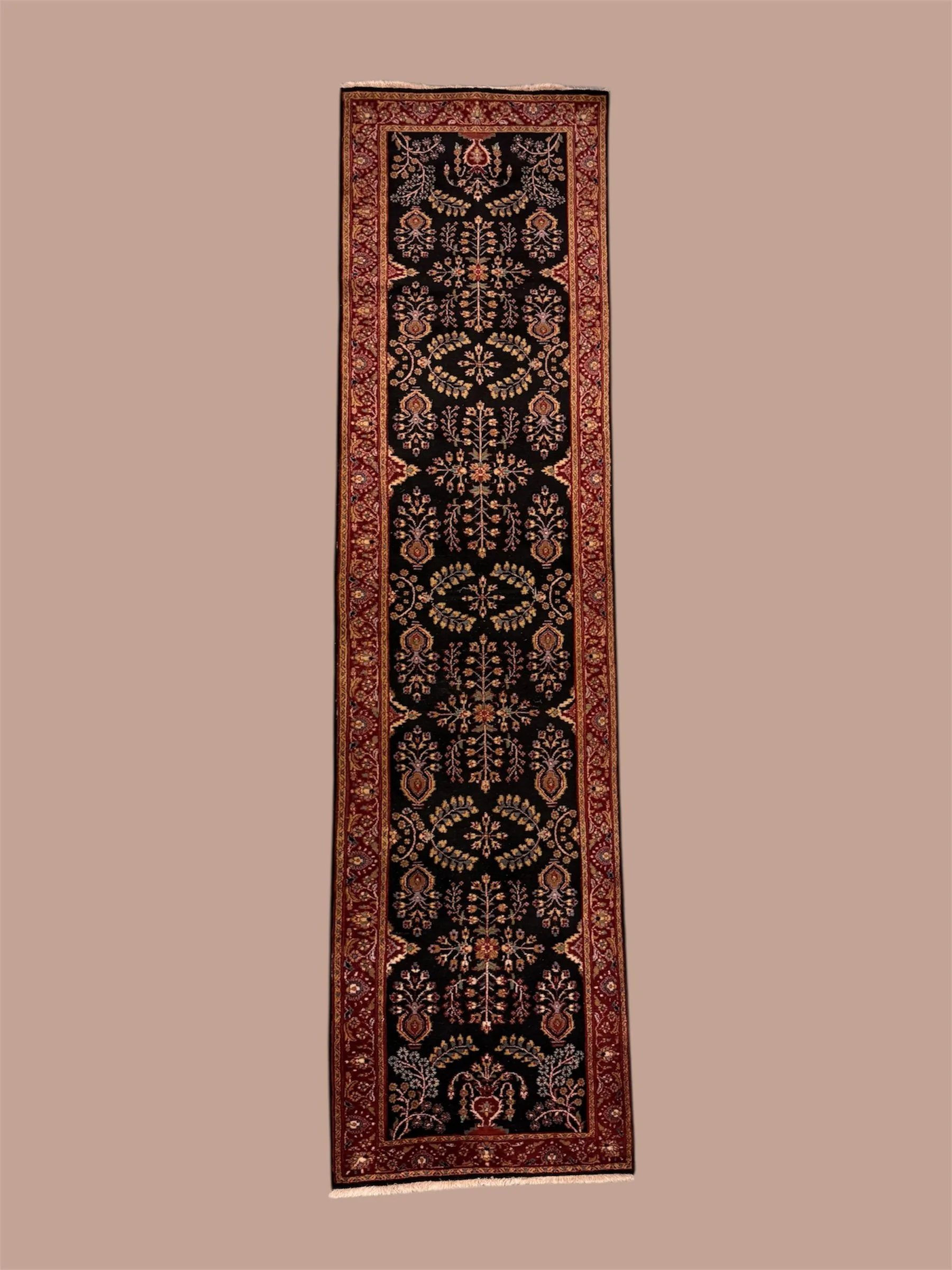 A long, narrow, ornate black area rug with intricate floral patterns in beige, red, and gold, bordered by a red and gold decorative border, placed on a light brown surface.