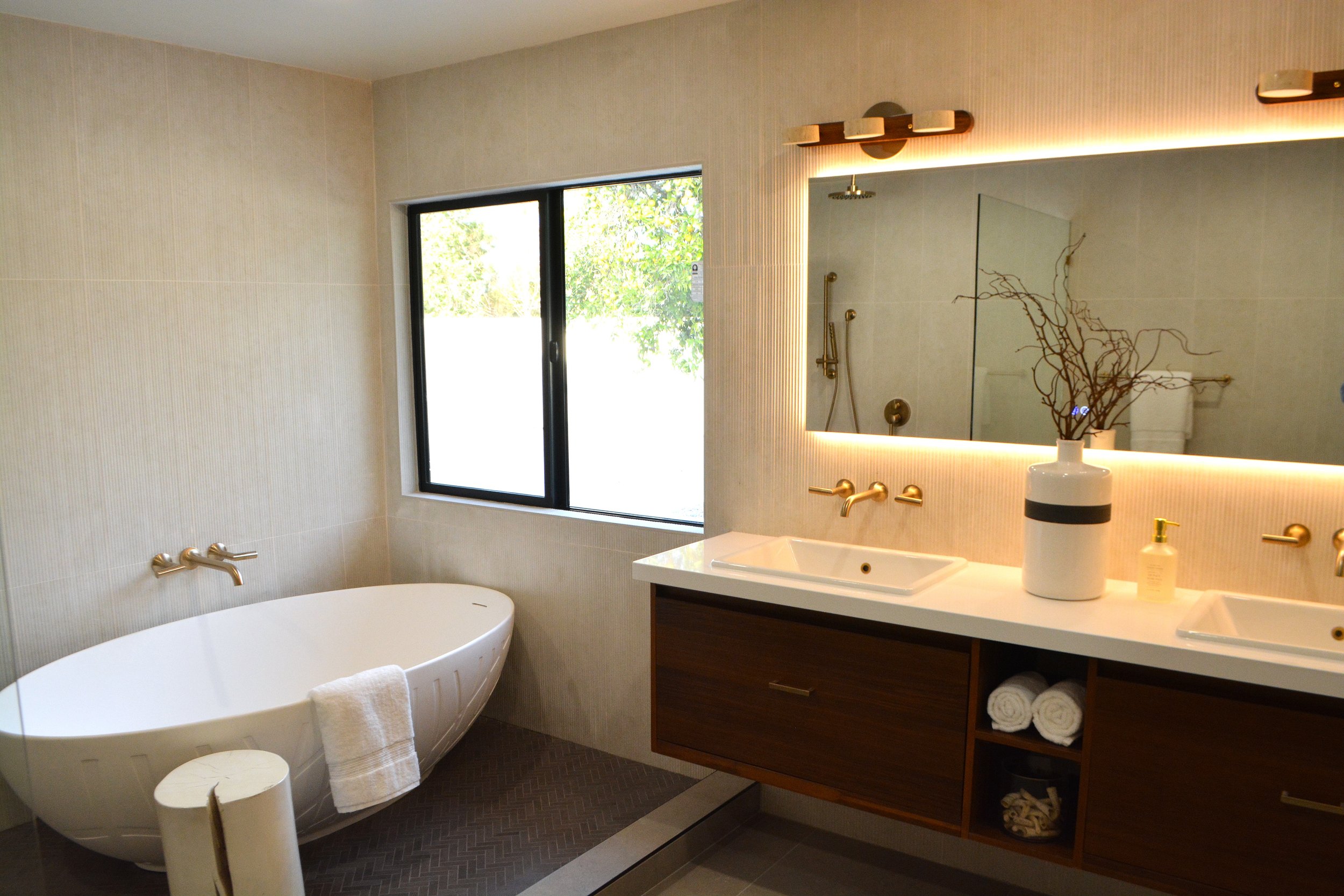 A modern bathroom with a large window, a white freestanding bathtub, a bathroom vanity with a sink, a large mirror with LED lighting, and decorative items like a vase with branches, soap, and towels.