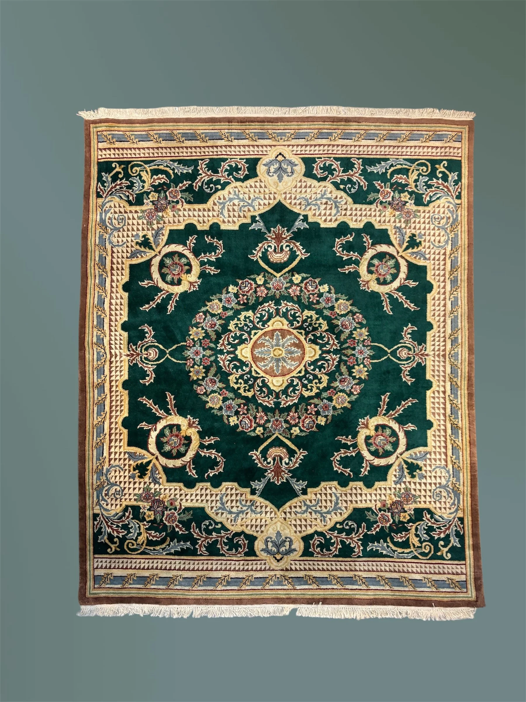 A detailed Persian-style area rug with floral and geometric patterns in green, beige, pink, and blue, with fringed edges on a plain grey background.