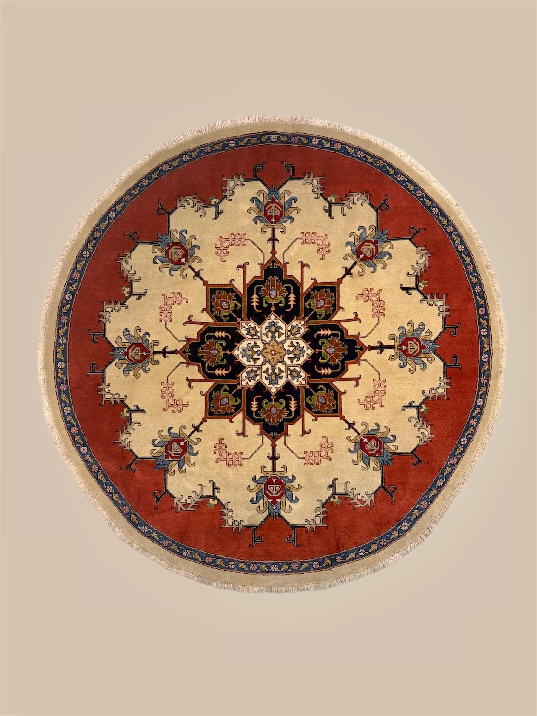 A round, decorative rug with intricate patterns in colors including red, black, beige, blue, and white, featuring symmetrical geometric and floral motifs.
