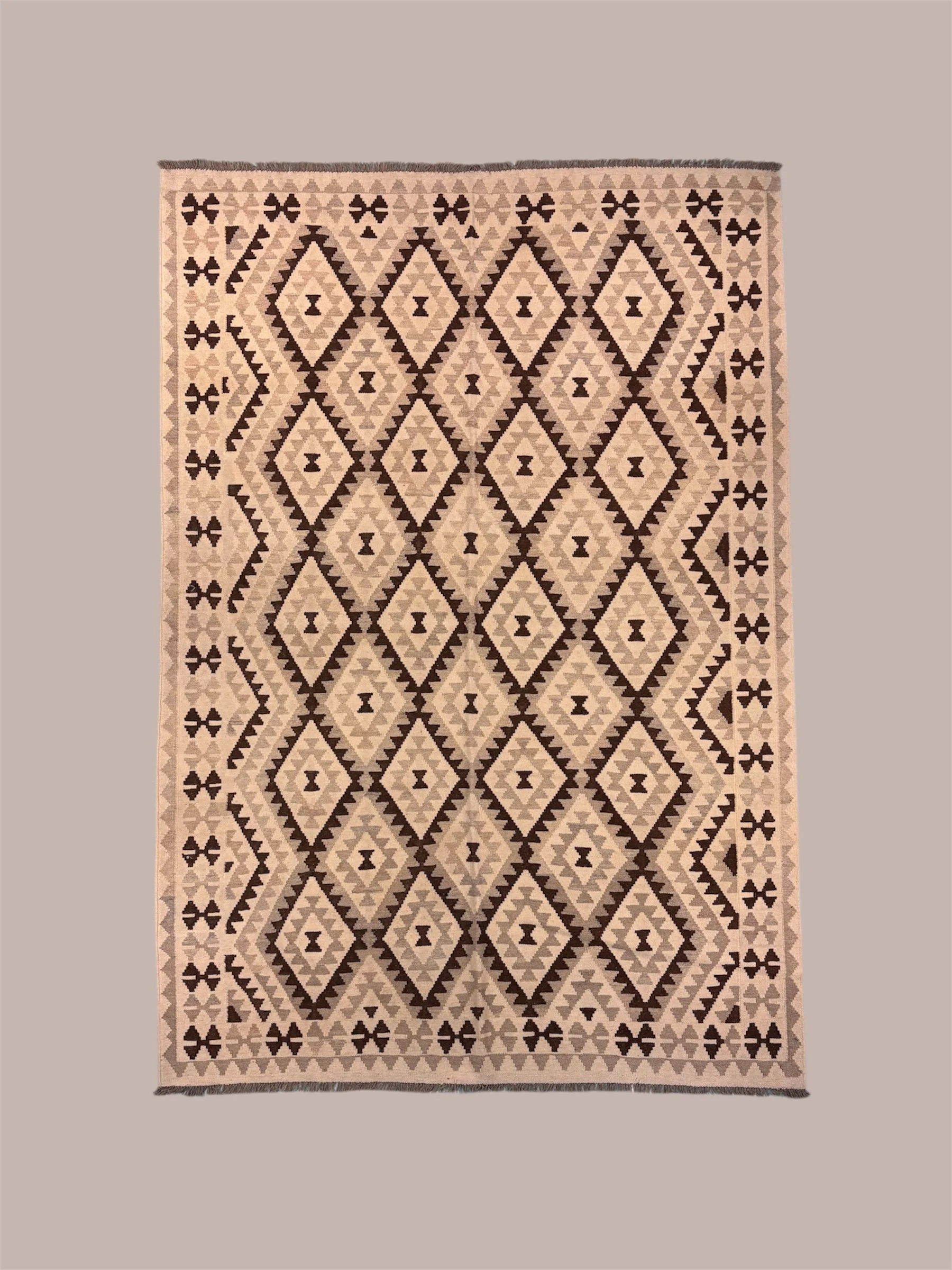 A patterned rug with a geometric diamond and cross design in shades of beige, brown, and cream, with fringes at the top and bottom edges.