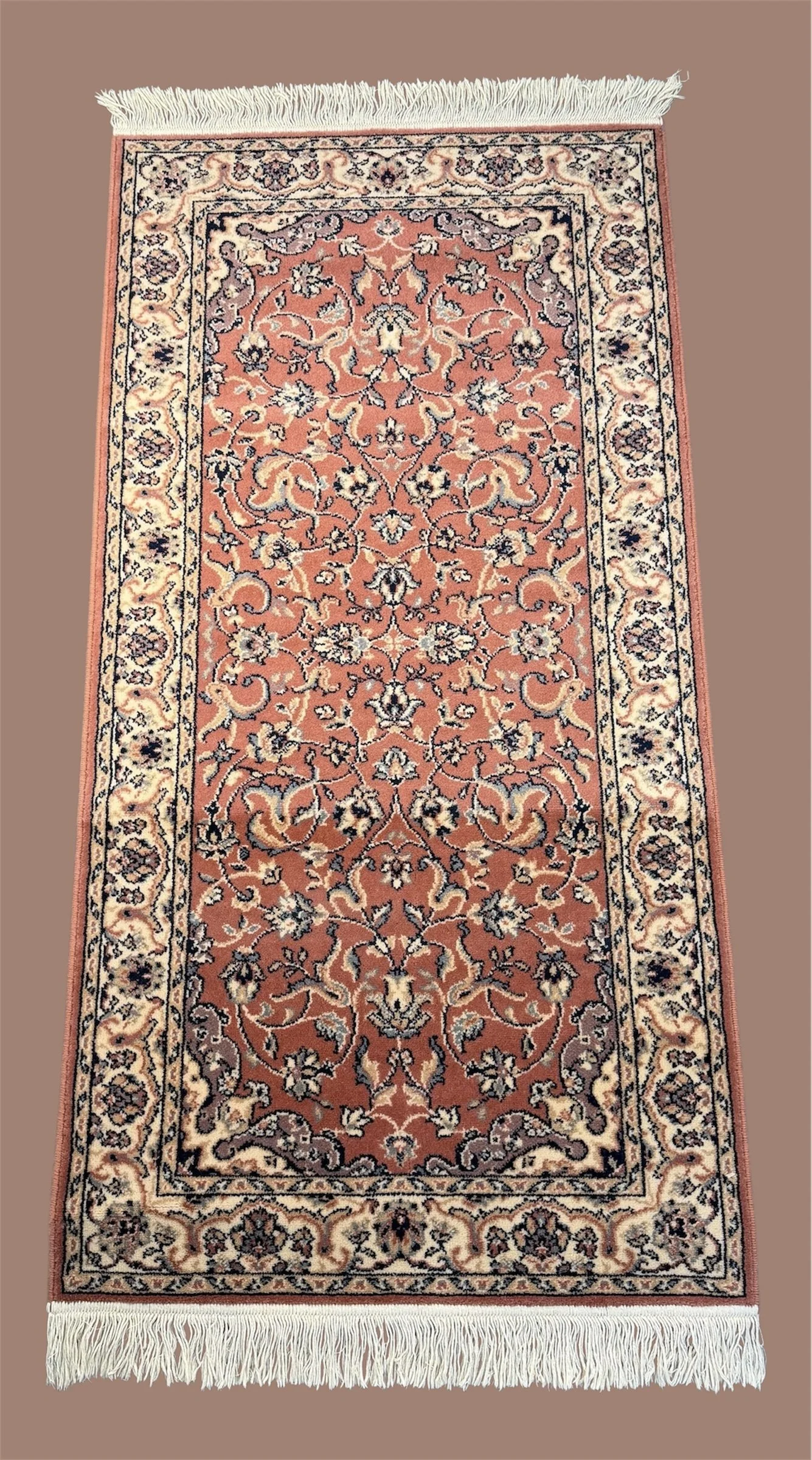 An intricately patterned, rectangular Persian-style rug with floral and vine motifs in shades of cream, black, beige, and rust, featuring a fringed border on both ends, placed on a pinkish-brown floor.