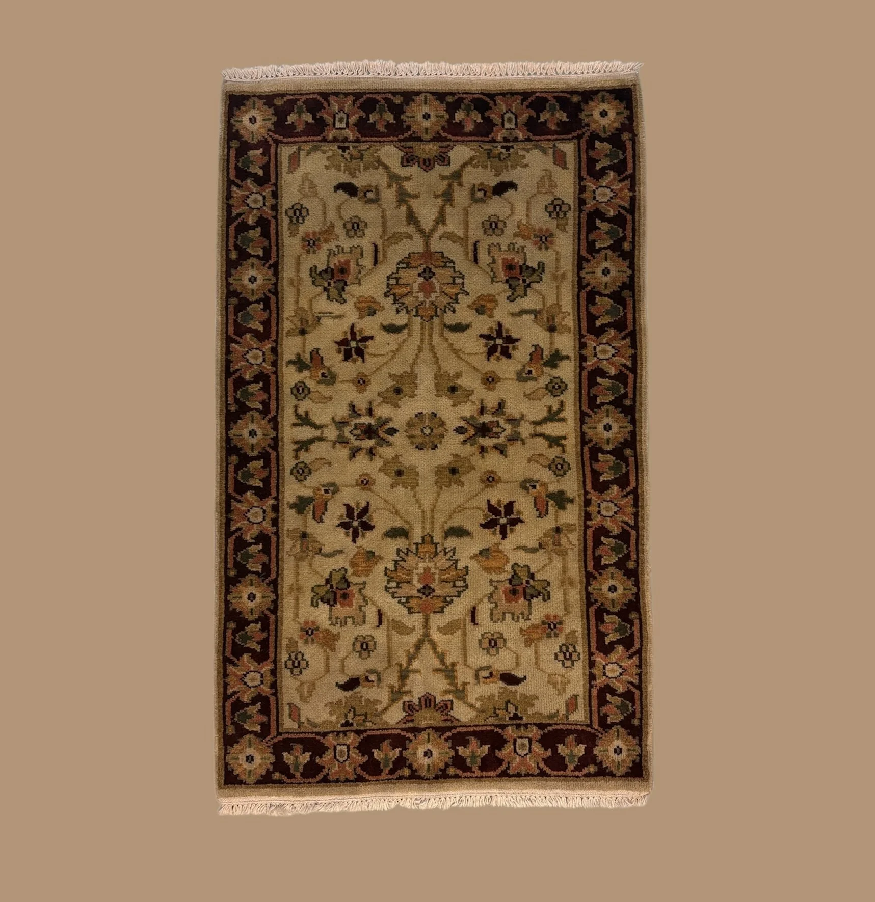 A rectangular area rug with a beige background and a floral and vine pattern in shades of orange, green, and dark brown, bordered by a dark brown and floral design with fringe on at least one end.