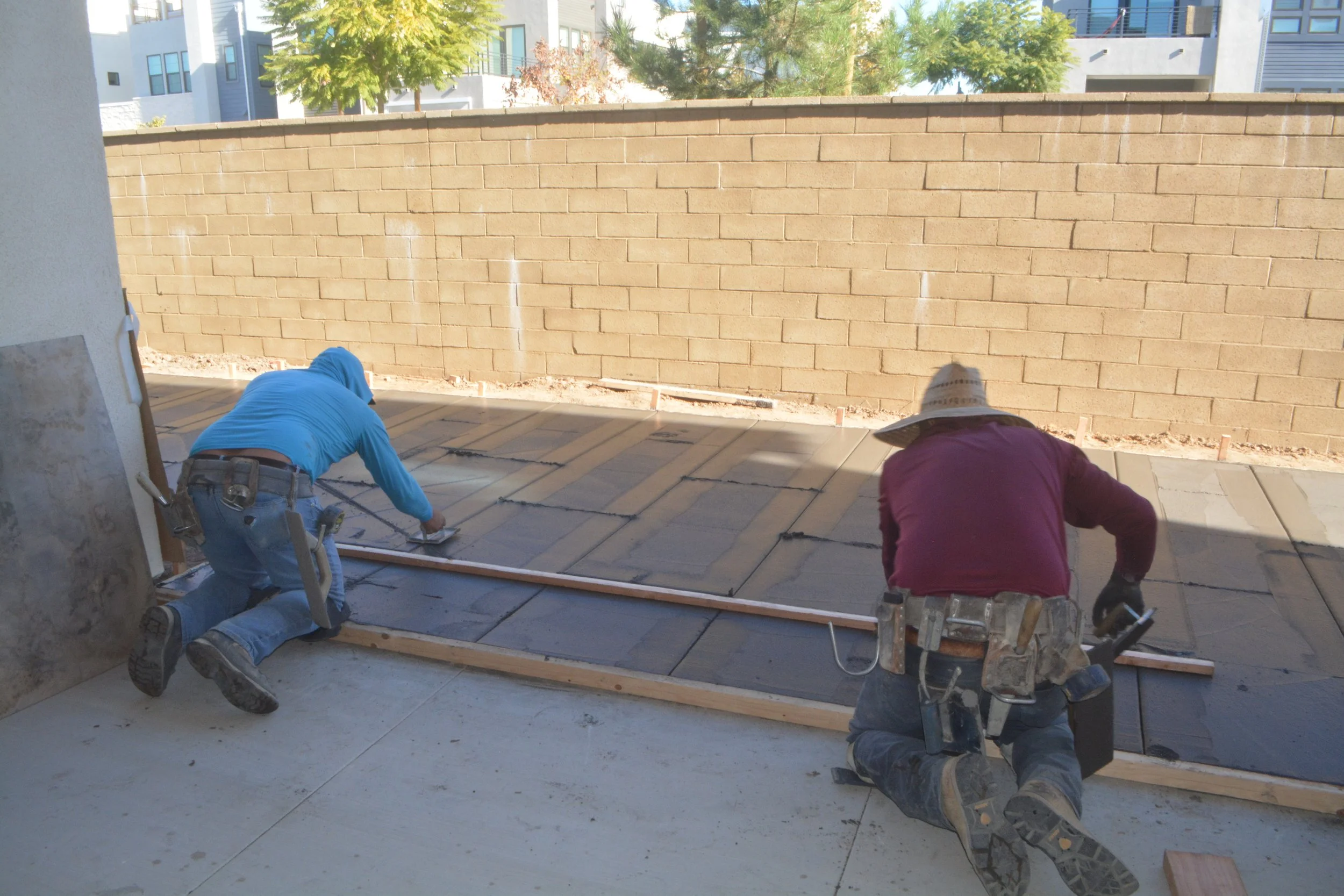 Two construction workers kneeling on a concrete patio, laying large paving stones in front of a brick wall during daytime.