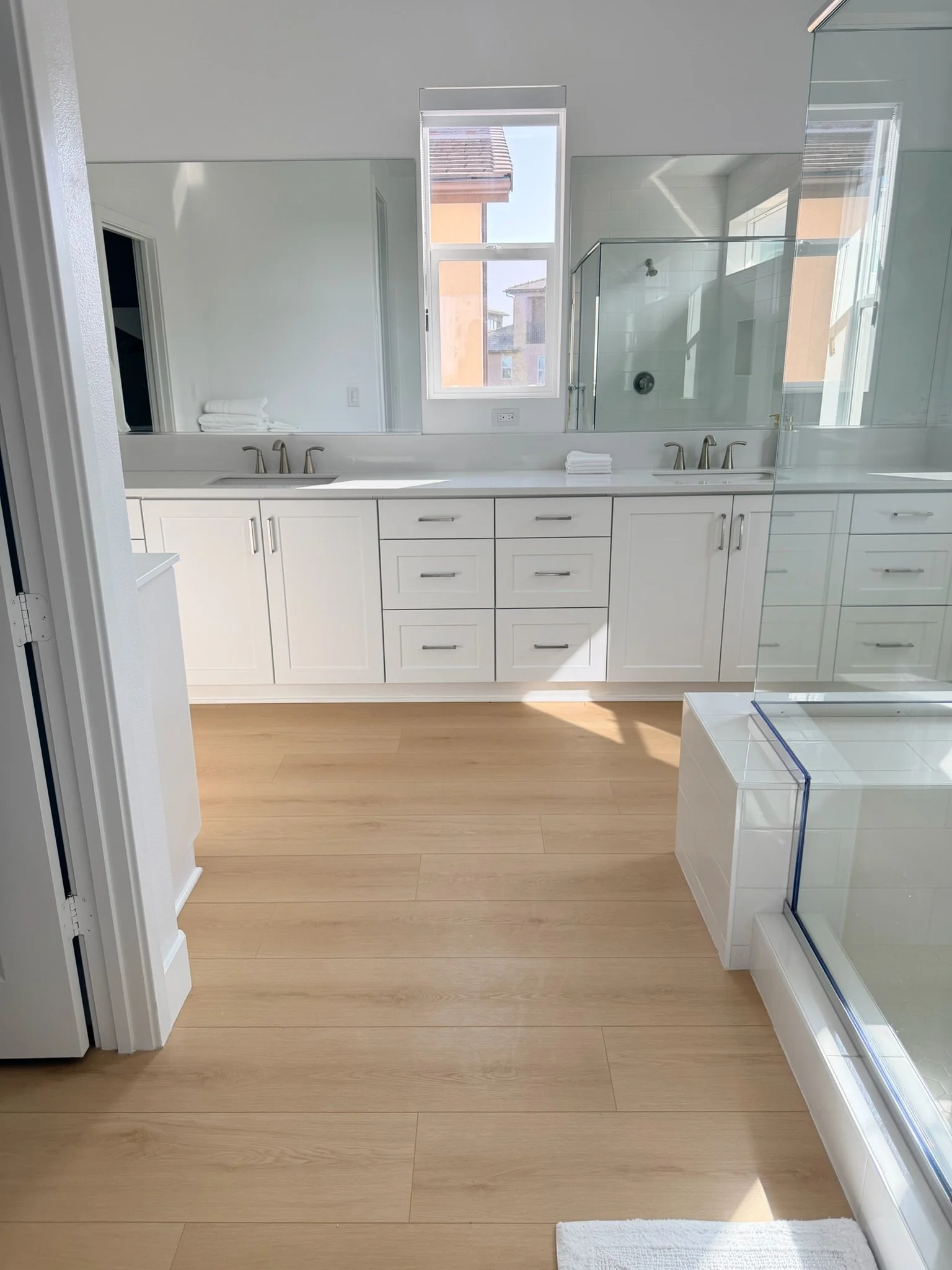 Bright modern bathroom featuring a double vanity with white cabinets and two sinks, a large mirror, a window with a view of neighboring rooftops, a glass-enclosed shower, and light wood flooring.
