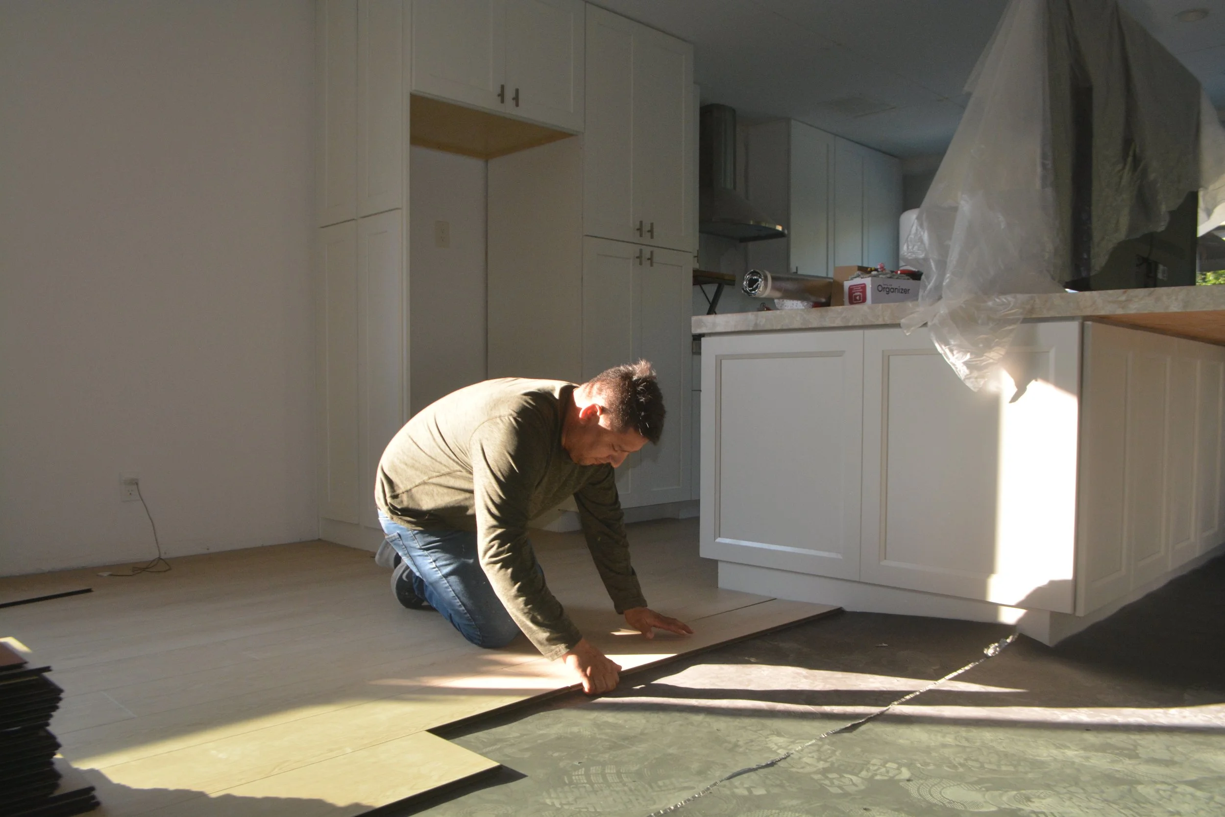 A man kneeling on the floor measuring and marking a section of new flooring in a kitchen with white cabinets and countertops. Sunlight is casting shadows on the floor.