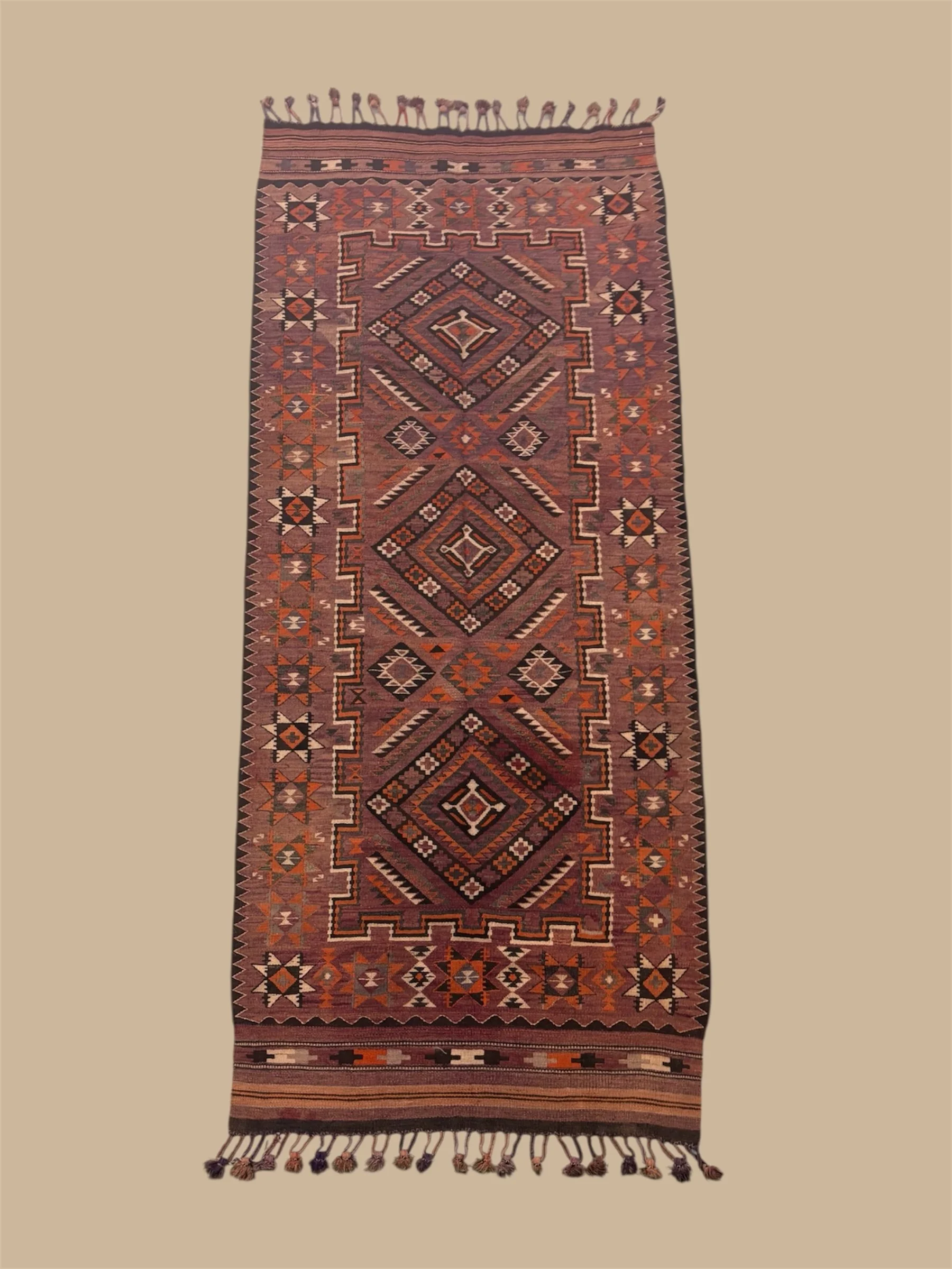 A long, narrow woven rug with geometric patterns in red, black, white, and beige, featuring star shapes and diamond motifs, with fringed tassels at the top and bottom.