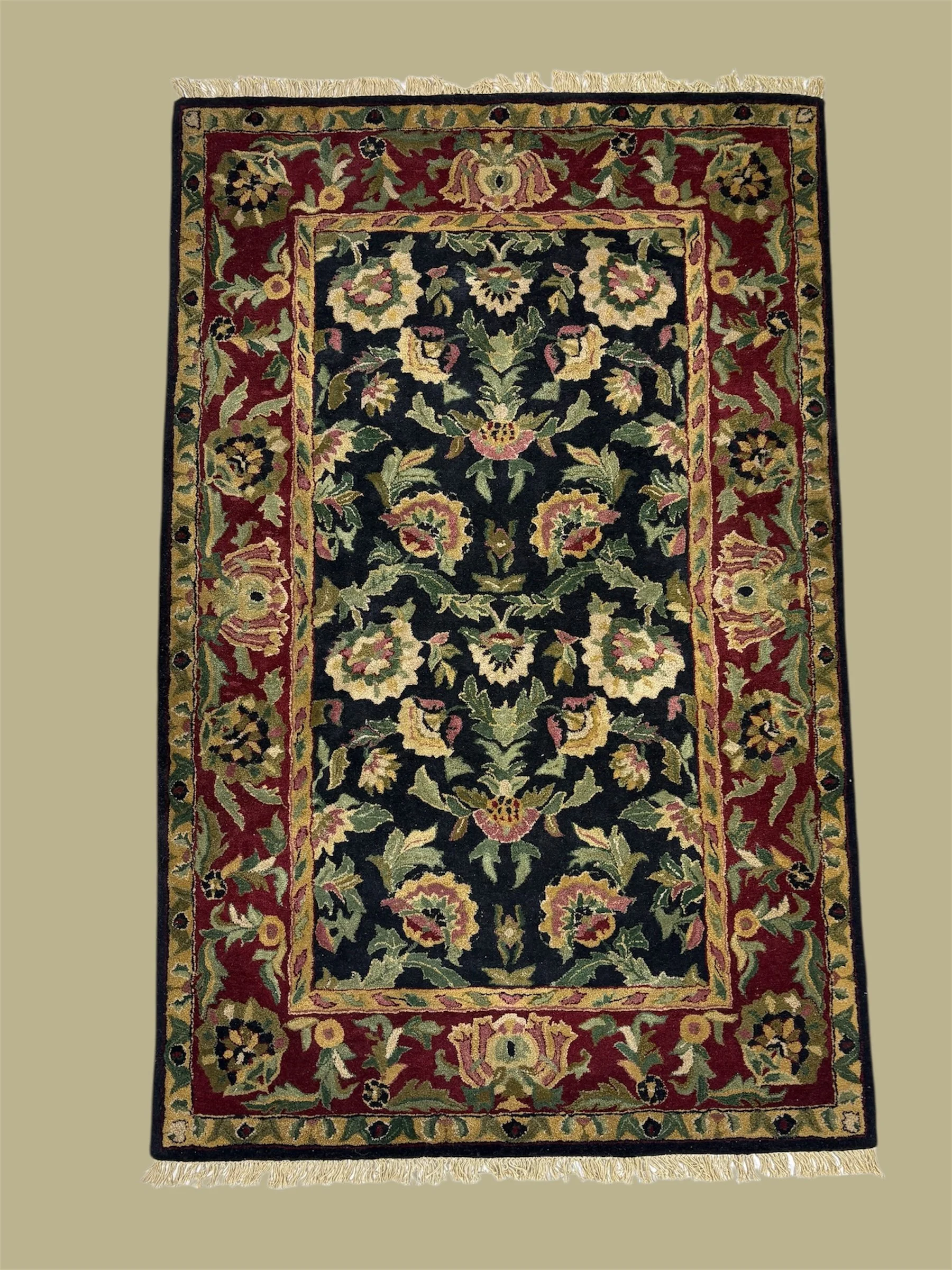 A traditional, intricately patterned area rug with floral motifs, featuring a central black field surrounded by a red and gold border, with fringes on both ends.