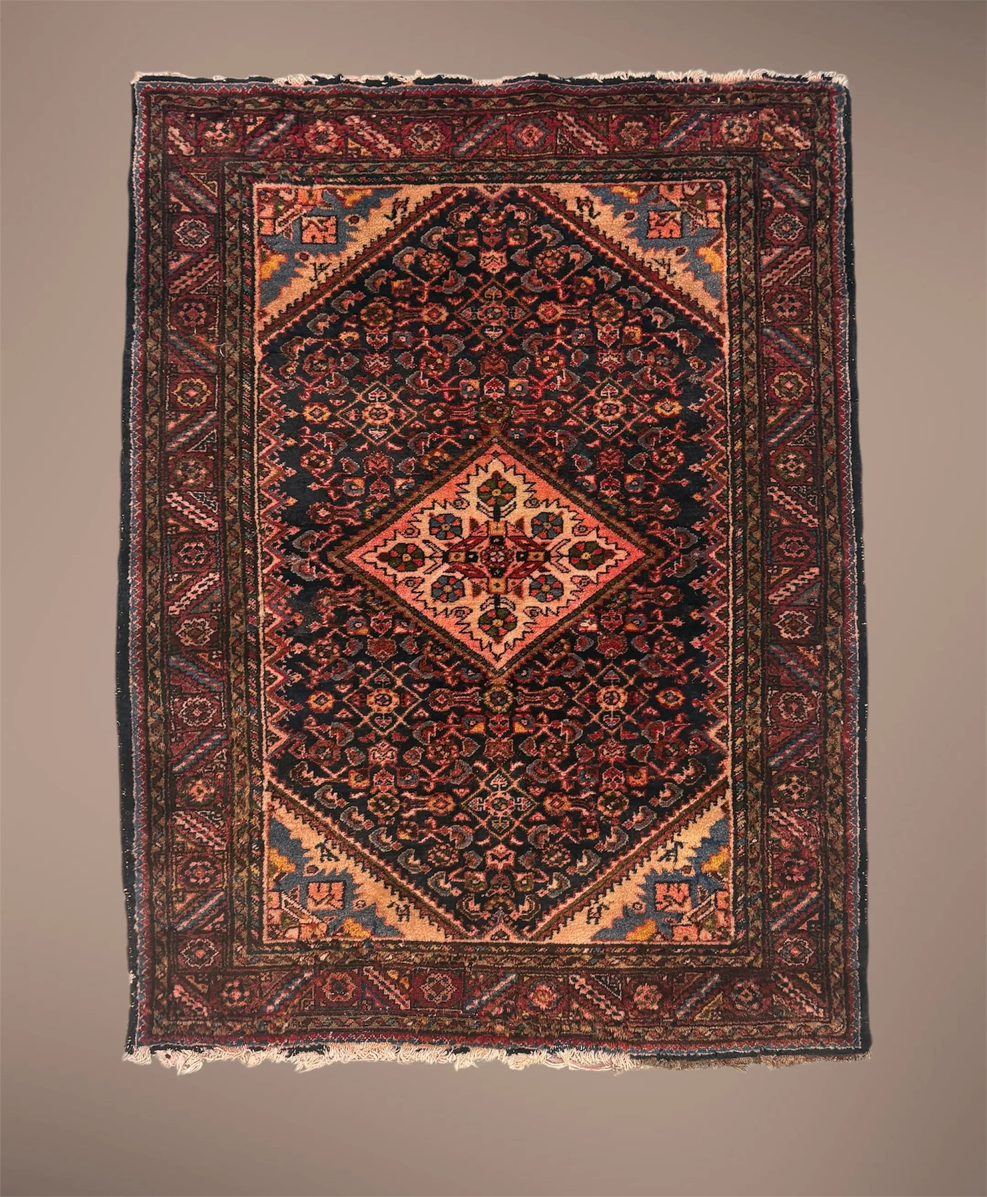 A traditional Persian-style rug with intricate floral and geometric patterns in red, black, cream, and gold tones, laying flat on a gray surface, with fringed edges visible at the top and bottom.