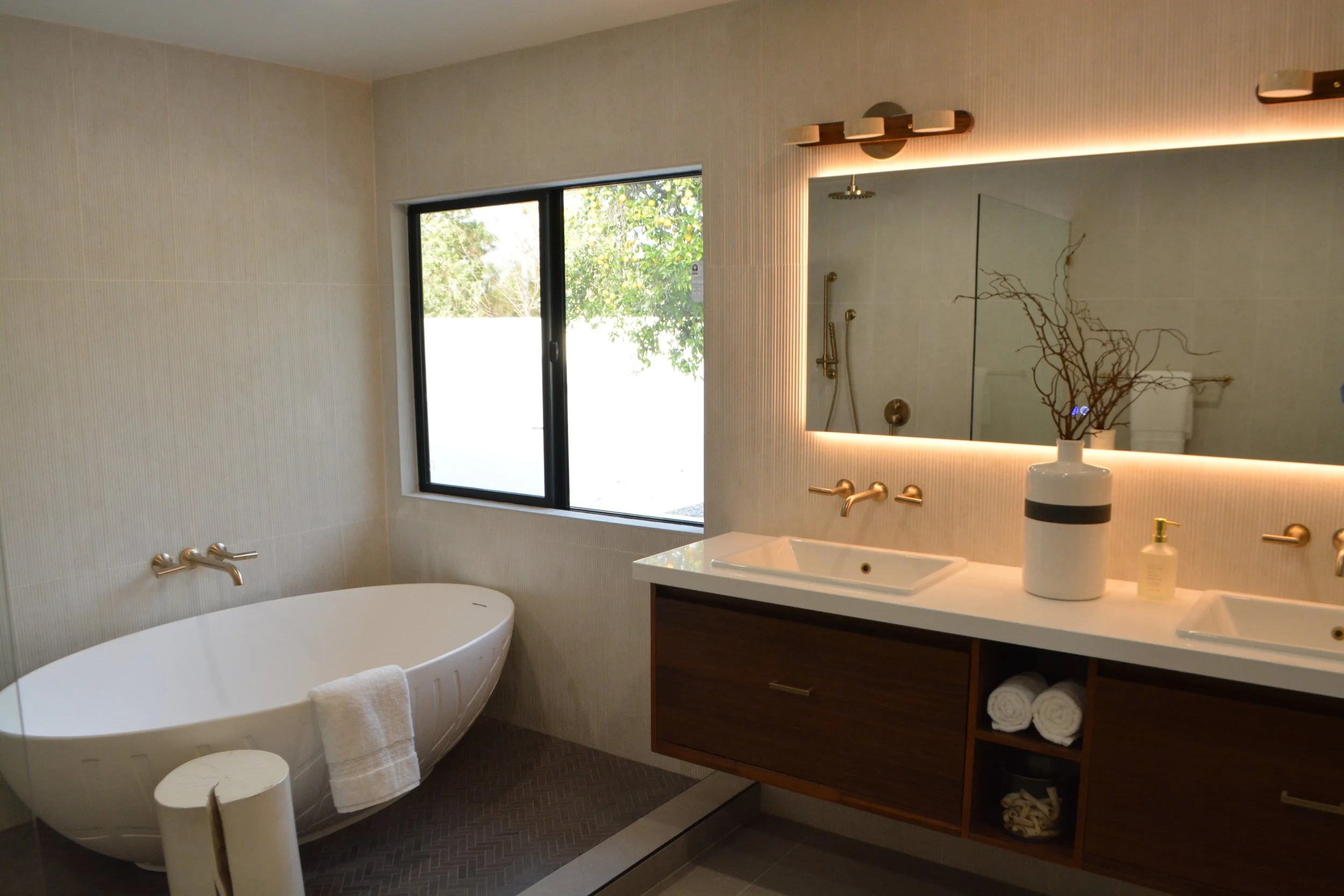 Modern bathroom with a freestanding bathtub, a large mirror with backlighting, a window showing greenery outside, and wooden cabinetry with rolled towels.