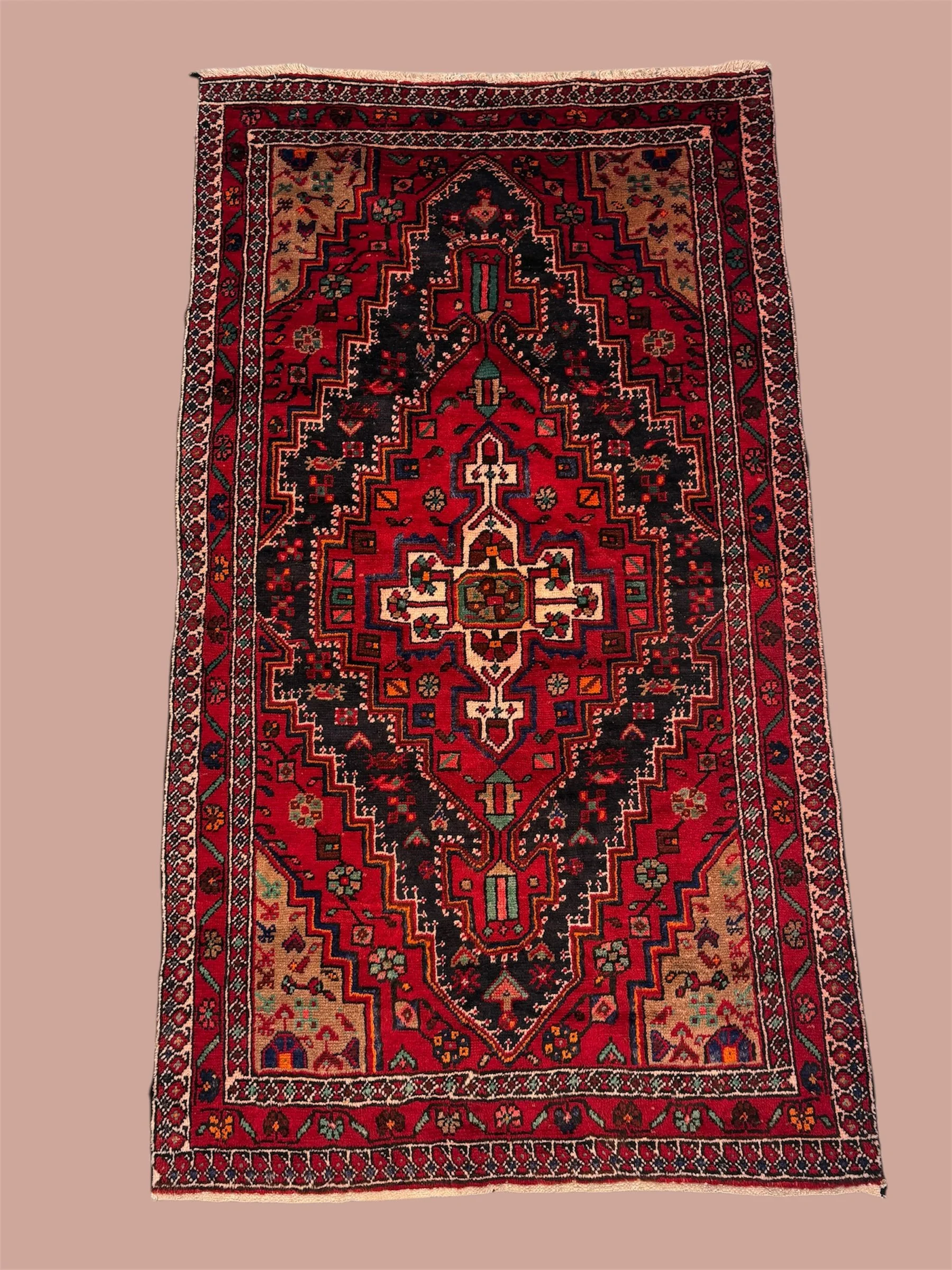A traditional, colorful patterned rug with a central diamond shape surrounded by floral and geometric designs, featuring red, black, cream, and green colors.