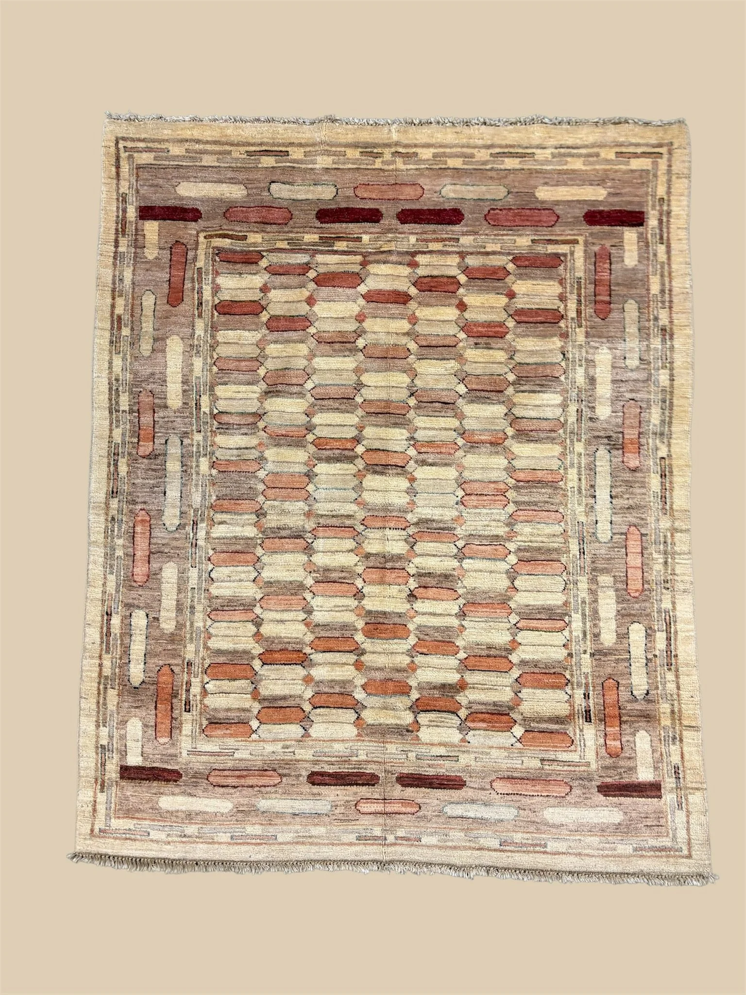 An area rug with a geometric pattern of rectangles and elongated shapes in shades of beige, red, and brown, with a detailed border and fringe at one end.