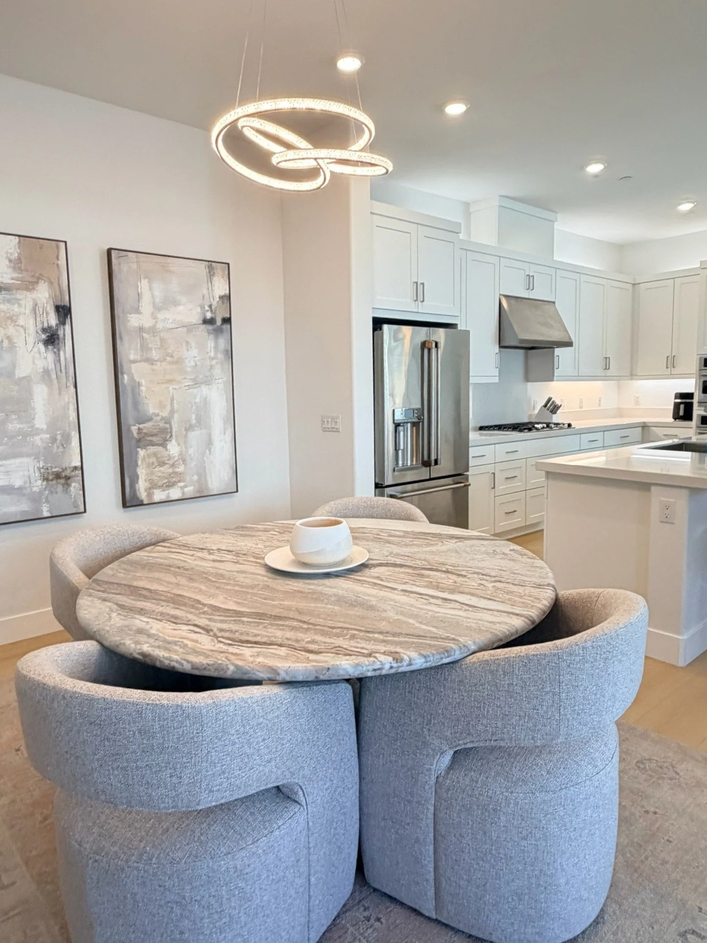 A modern kitchen with white cabinets, stainless steel refrigerator, and a round marble dining table with plush chairs, decorated with abstract wall art and a contemporary ceiling light fixture.