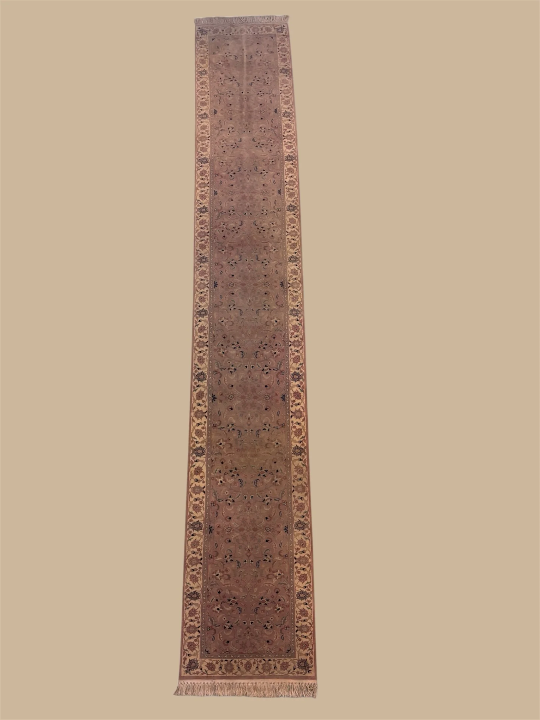 Long, ornate runner rug with intricate floral and geometric patterns in shades of beige, pink, and dark blue, with fringed ends.