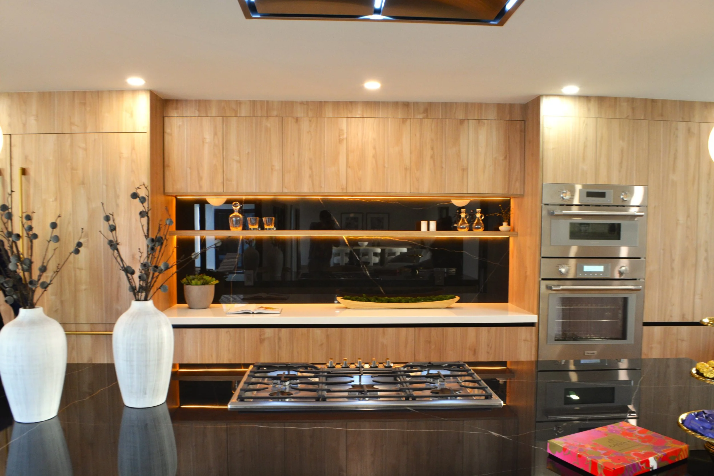 Modern kitchen with light wood cabinetry, black countertop, built-in oven, and gas stove. Decor includes three white vases with black branches, a potted plant, and glasses with decanters. Recessed ceiling lights.