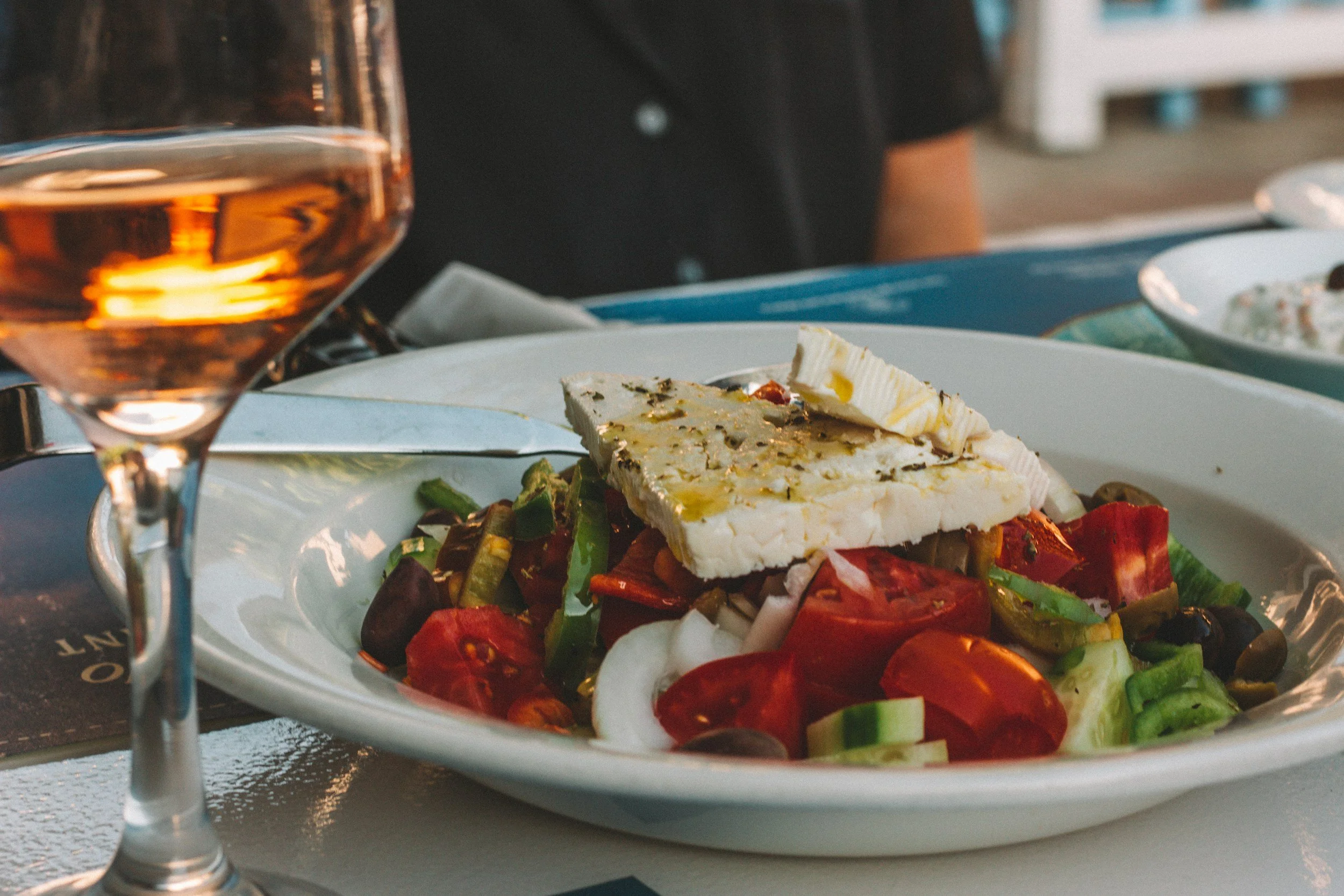 A plate of Greek salad with tomatoes, cucumbers, red onions, olives, and feta cheese, with a glass of rose wine beside it.