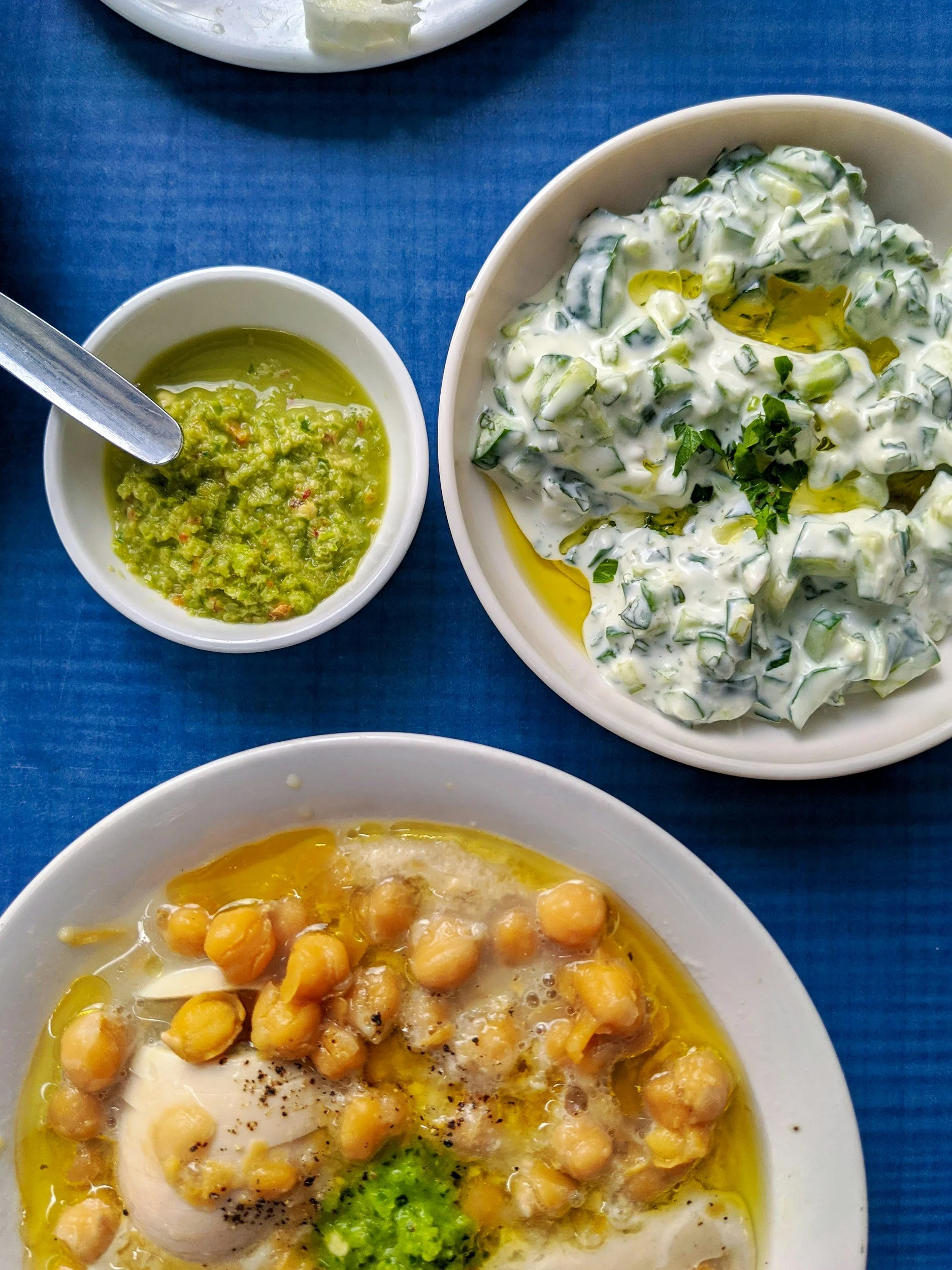 A plate of hummus topped with whole chickpeas, olive oil, and black pepper, with a side of chopped parsley. A bowl of creamy cucumber salad with yogurt, cucumber, and herbs, drizzled with olive oil. A small bowl of green dipping sauce, possibly guaca