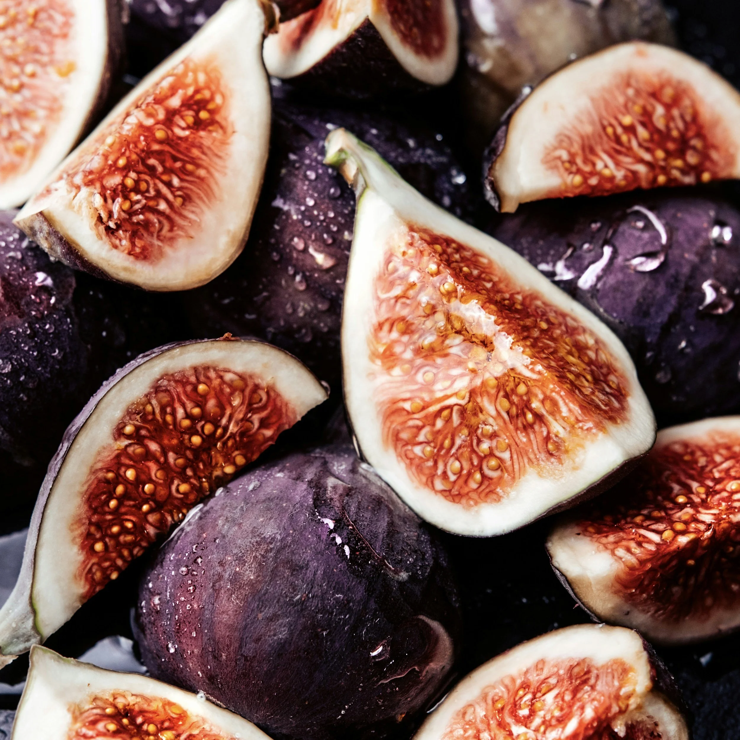 Close-up of fresh figs, some sliced open, showing their red, seed-filled interior, with droplets of water on the dark purple skins.