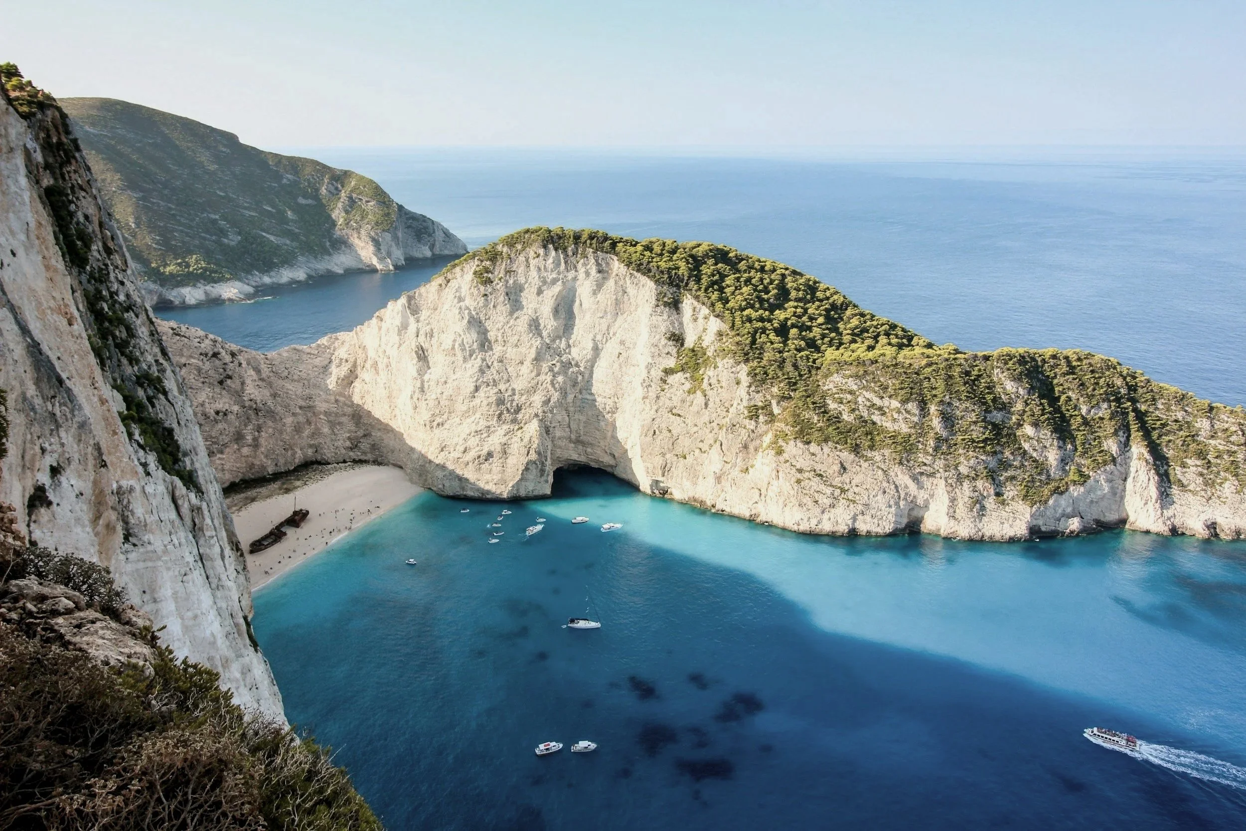 Aerial view of Navagio Beach with a shipwreck, white sandy beach, turquoise water, and steep cliffs with green vegetation.