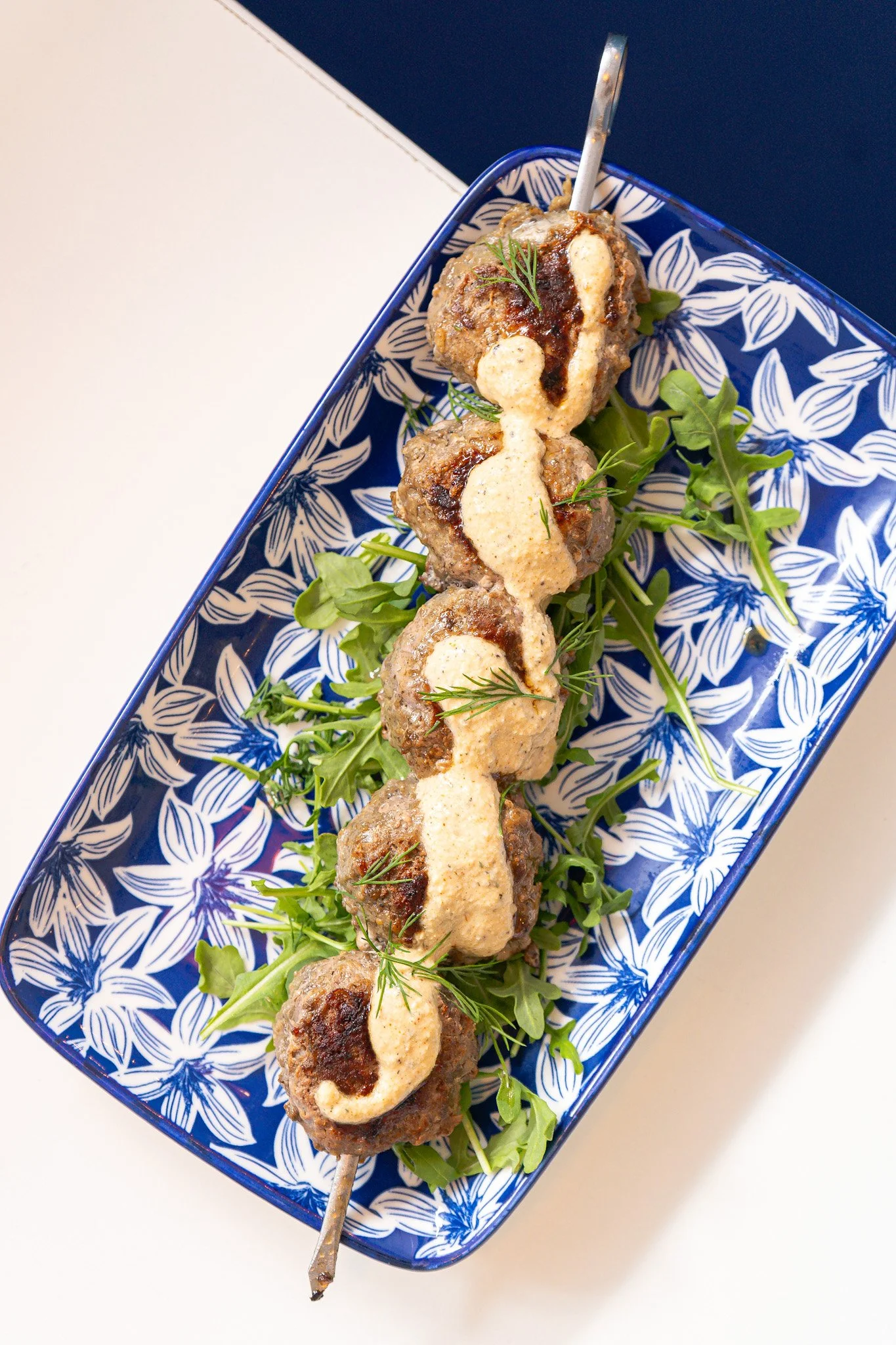 Skewered meatballs garnished with green herbs, served on a blue and white patterned rectangular dish with fresh greens.