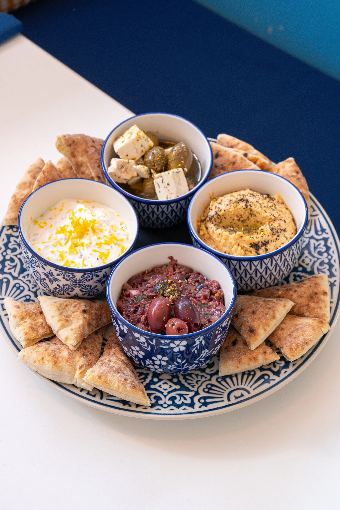 A large decorative plate filled with small bowls of various dips and spreads surrounding pita bread triangles.