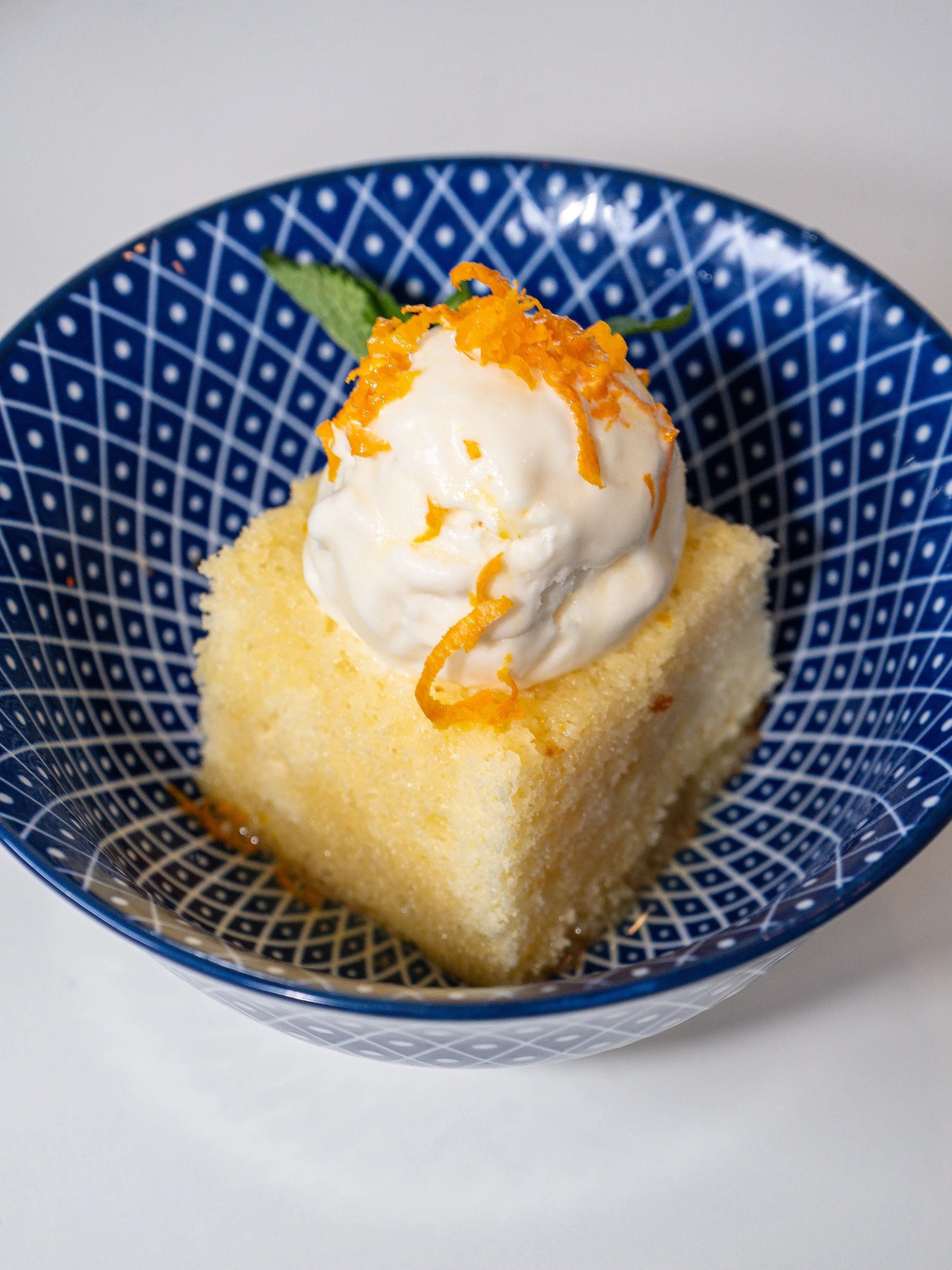 A yellow cake with a scoop of vanilla ice cream on top, garnished with shredded orange zest and a mint leaf, served in a blue patterned bowl.