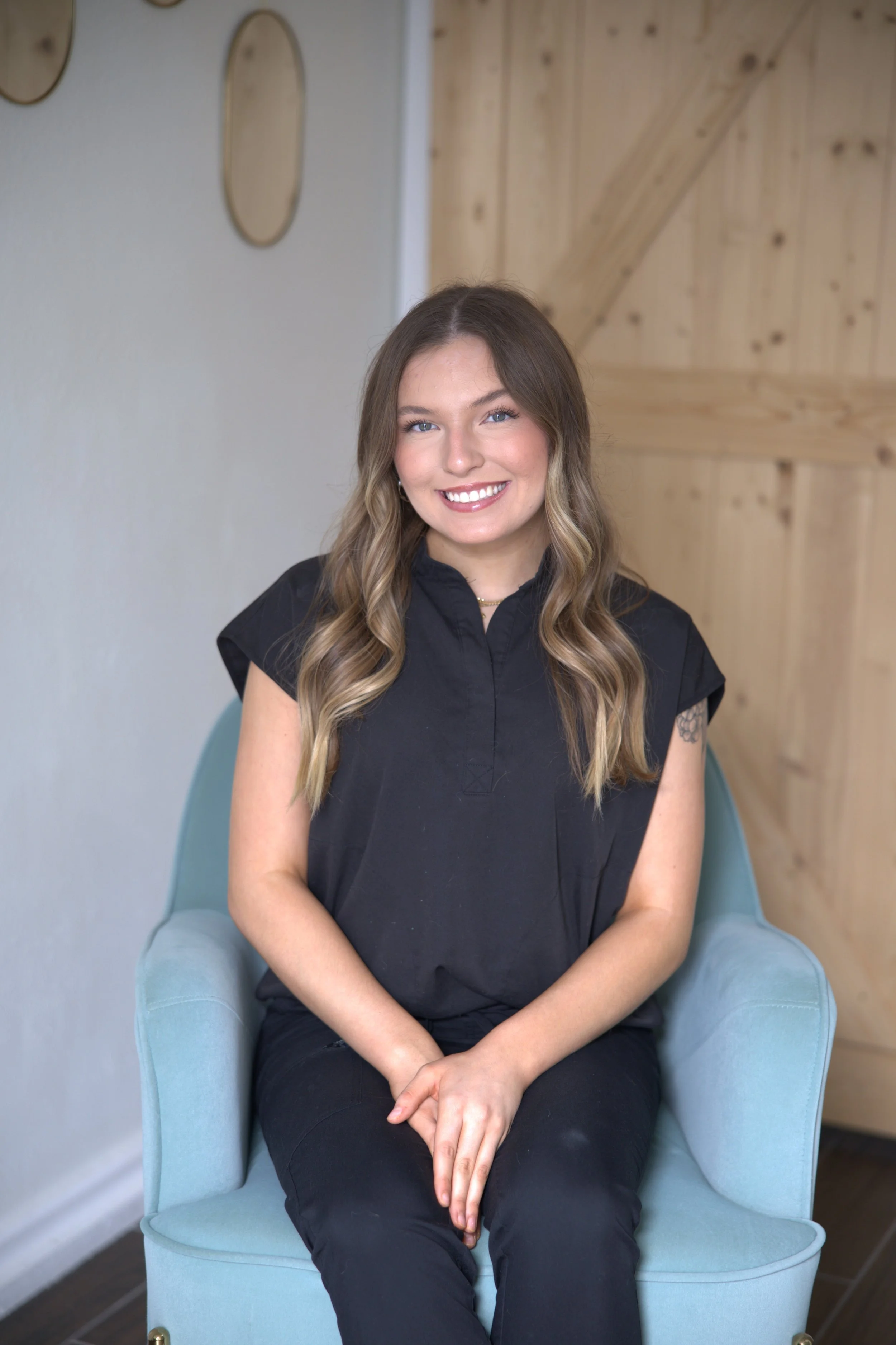 A young woman with long wavy hair and a smile, sitting in a light blue chair indoors with a wooden wall in the background.