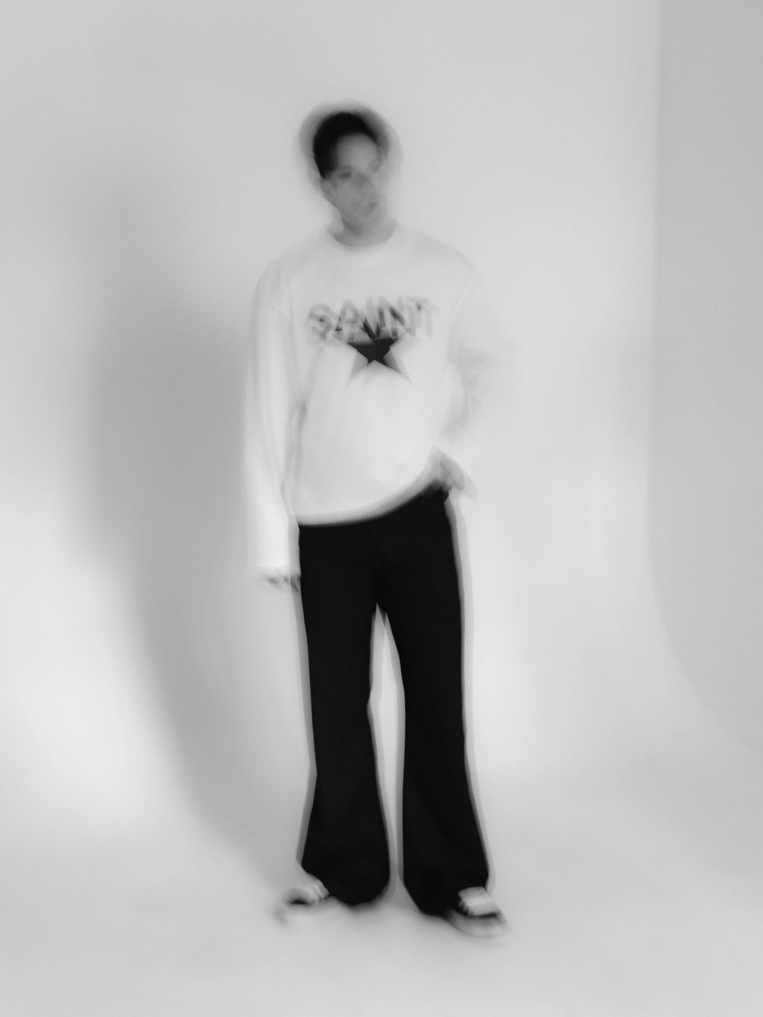 A person standing against a plain white background, wearing a white sweatshirt with 'SAINT' and a star printed on it, and dark pants.