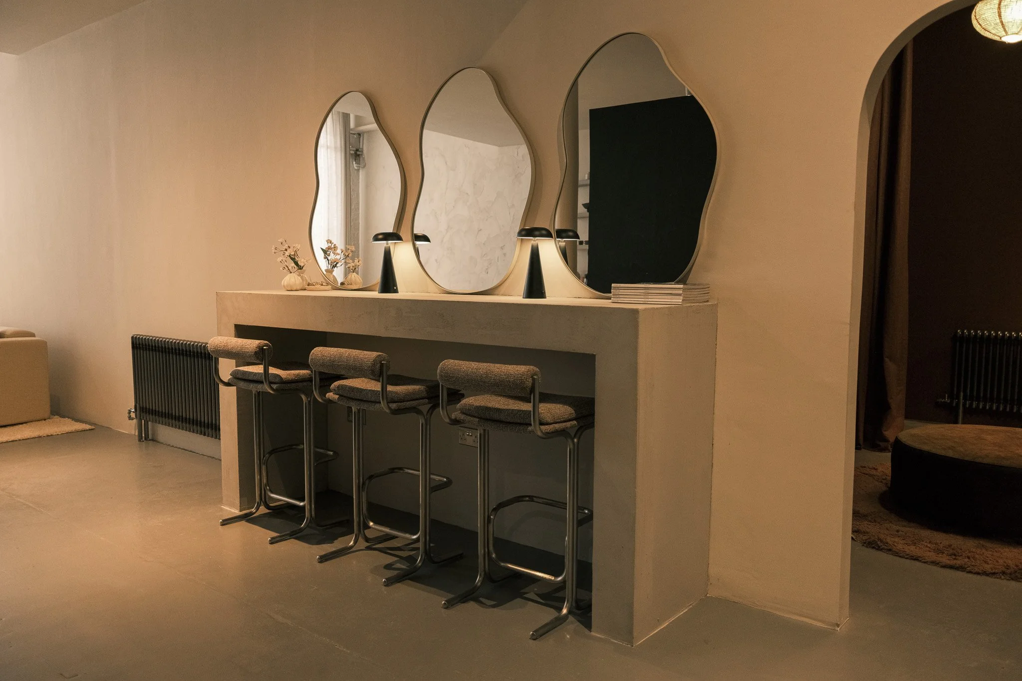 A modern bar area with a concrete counter, four barstools, three lamps, decorative vases with flowers, and three mirrors with irregular shapes on the wall.