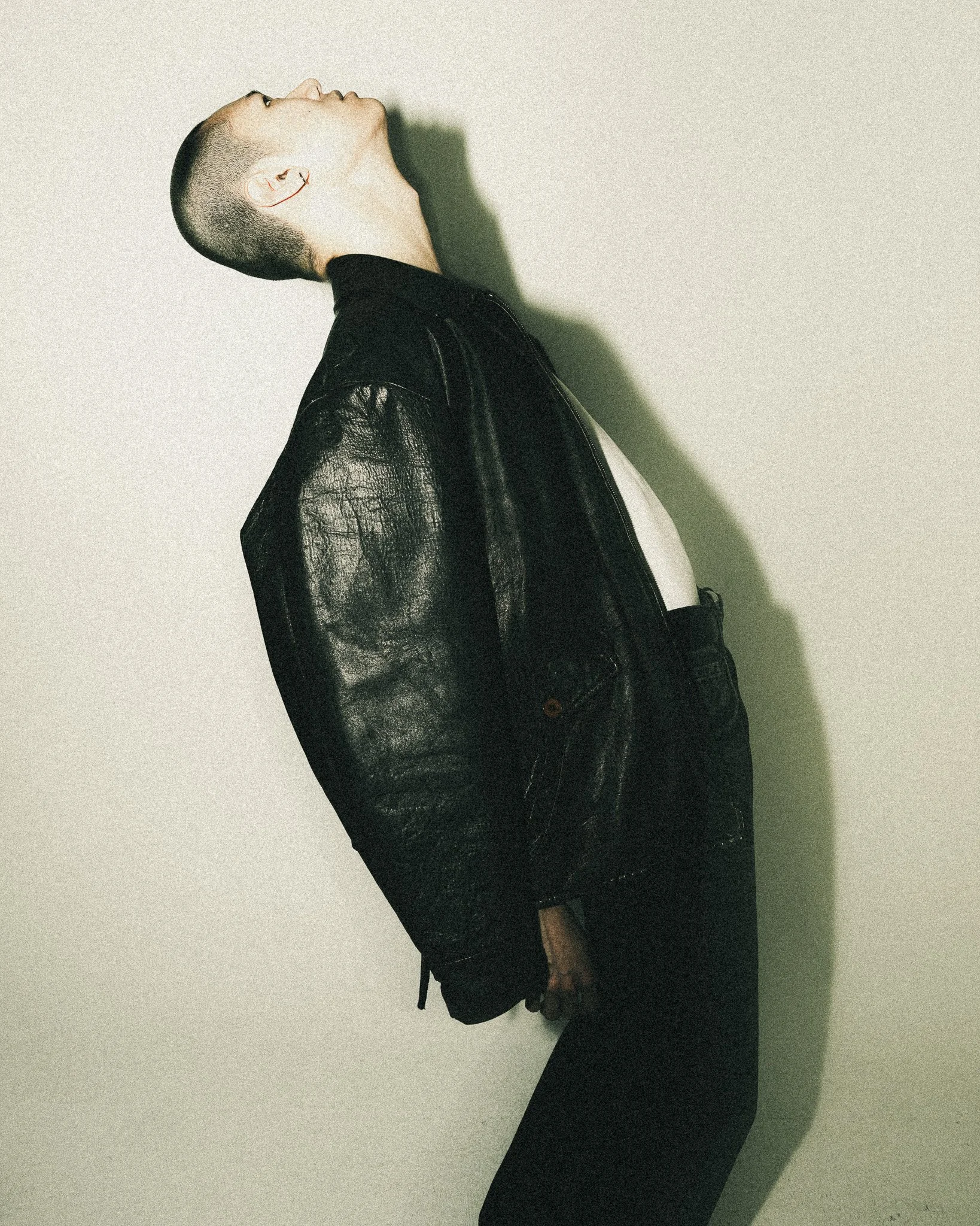Person with short hair wearing a black leather jacket, white shirt, and black pants, leaning back against a plain wall, with head tilted upward.
