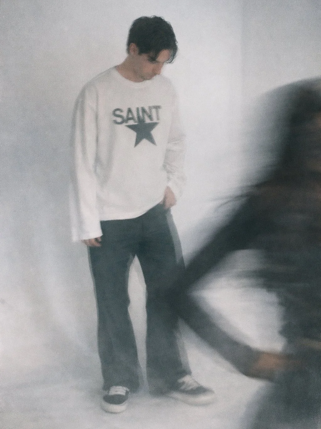 A young man with dark hair wearing a white Saint sweatshirt, dark pants, and sneakers standing against a plain wall, looking down.